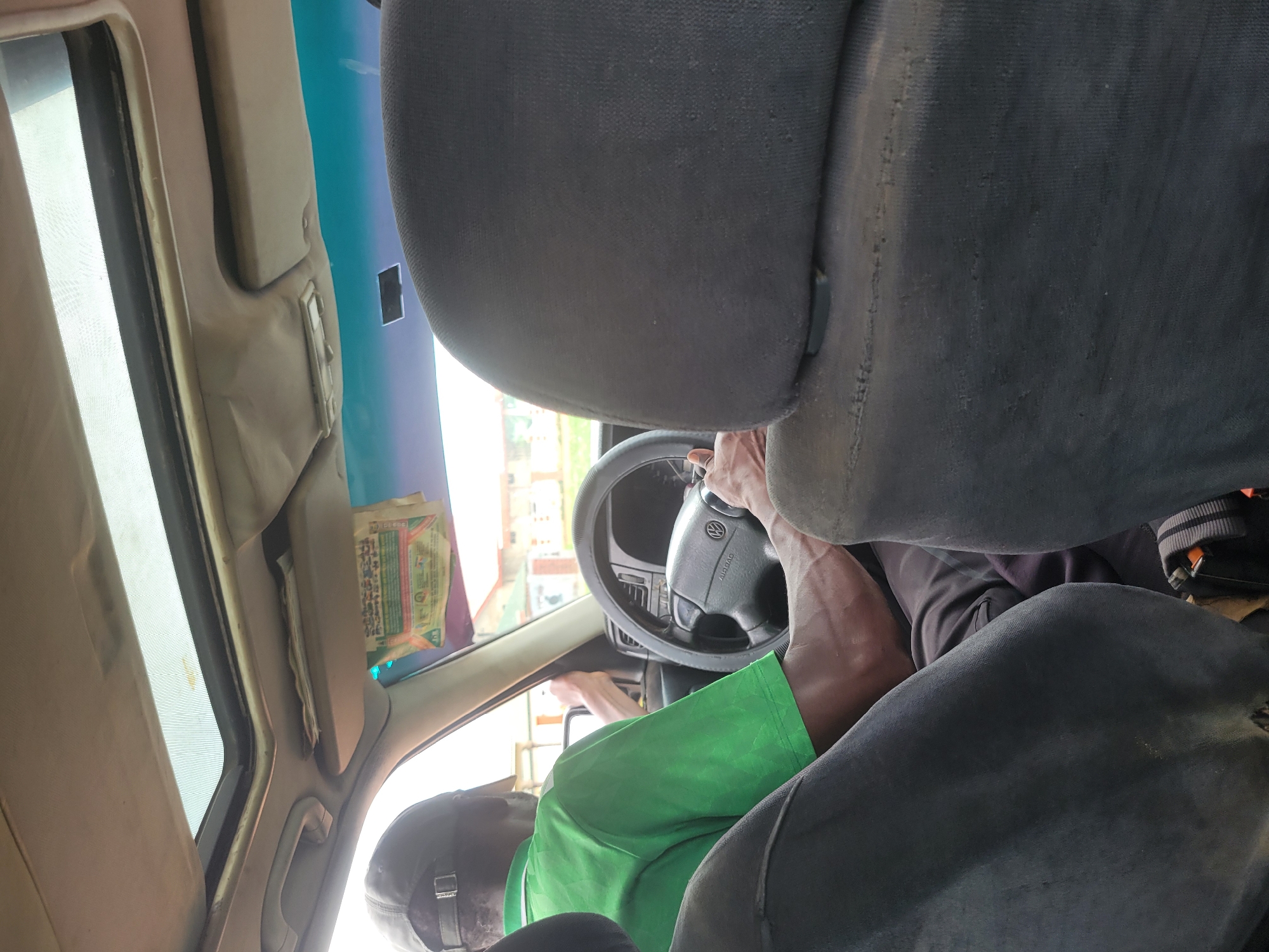 This is a first-person perspective inside a vehicle, likely a Volkswagen, in Bokkos, Nigeria. The driver, a Black male wearing a green t-shirt and a black baseball cap, is visible from the back and side as he steers the vehicle. His muscular arms are prominent as they grip the steering wheel. The interior of the car shows a sunroof, sun visors, and dark fabric seats. A piece of paper, possibly an advertisement or notice, is attached to the sun visor. The exterior view through the window reveals buildings and trees, suggesting an urban or semi-urban environment. The lighting suggests it is daytime.