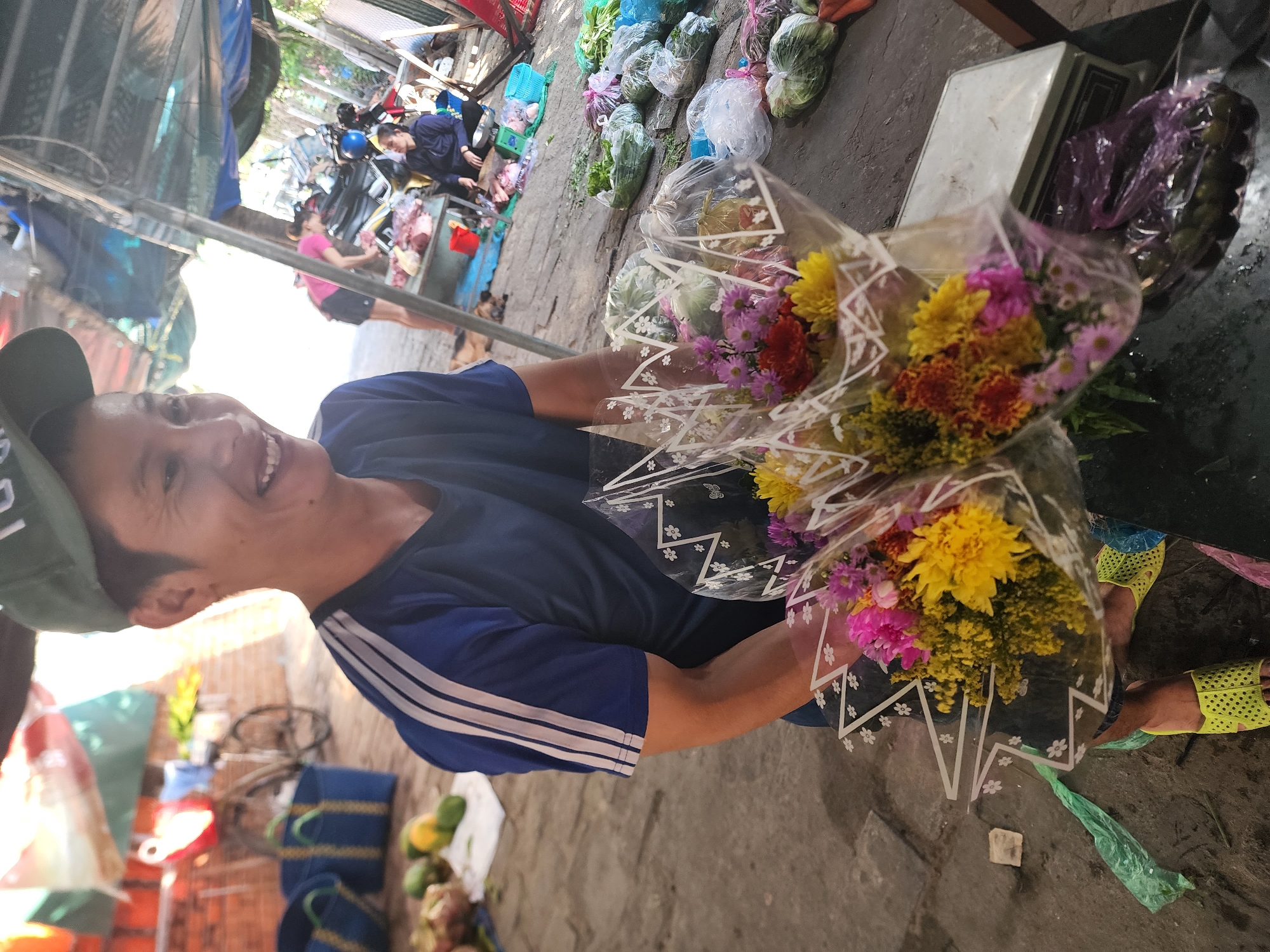 In Phu Thuy Ward, Vietnam, a person wearing a green baseball cap and a blue t-shirt with white stripes on the sleeves is holding two bouquets of flowers. The flowers are colorful, with yellow, pink, and orange blooms. The person is smiling, and their face is well-lit by sunlight. The background shows a market scene with various stalls and people. There are blue baskets, produce like fruits and vegetables, and a bicycle visible. A woman in a pink shirt is seen in the middle ground, and another woman is sitting behind a table in the background. The ground is made of concrete and dirt.