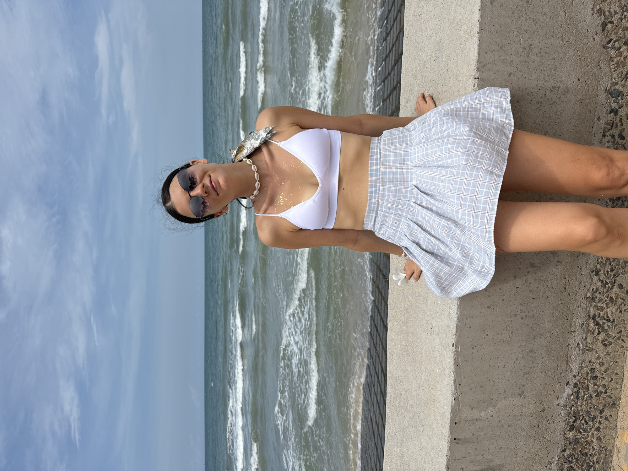 A woman wearing a white bikini top and a plaid skirt sits on a concrete ledge overlooking the ocean. A scarf is tied around her neck, and a small fish is draped over her shoulder. She wears sunglasses and a seashell necklace. The ocean behind her is rough with waves, and the sky is cloudy. The location is Mũi Né, Vietnam.