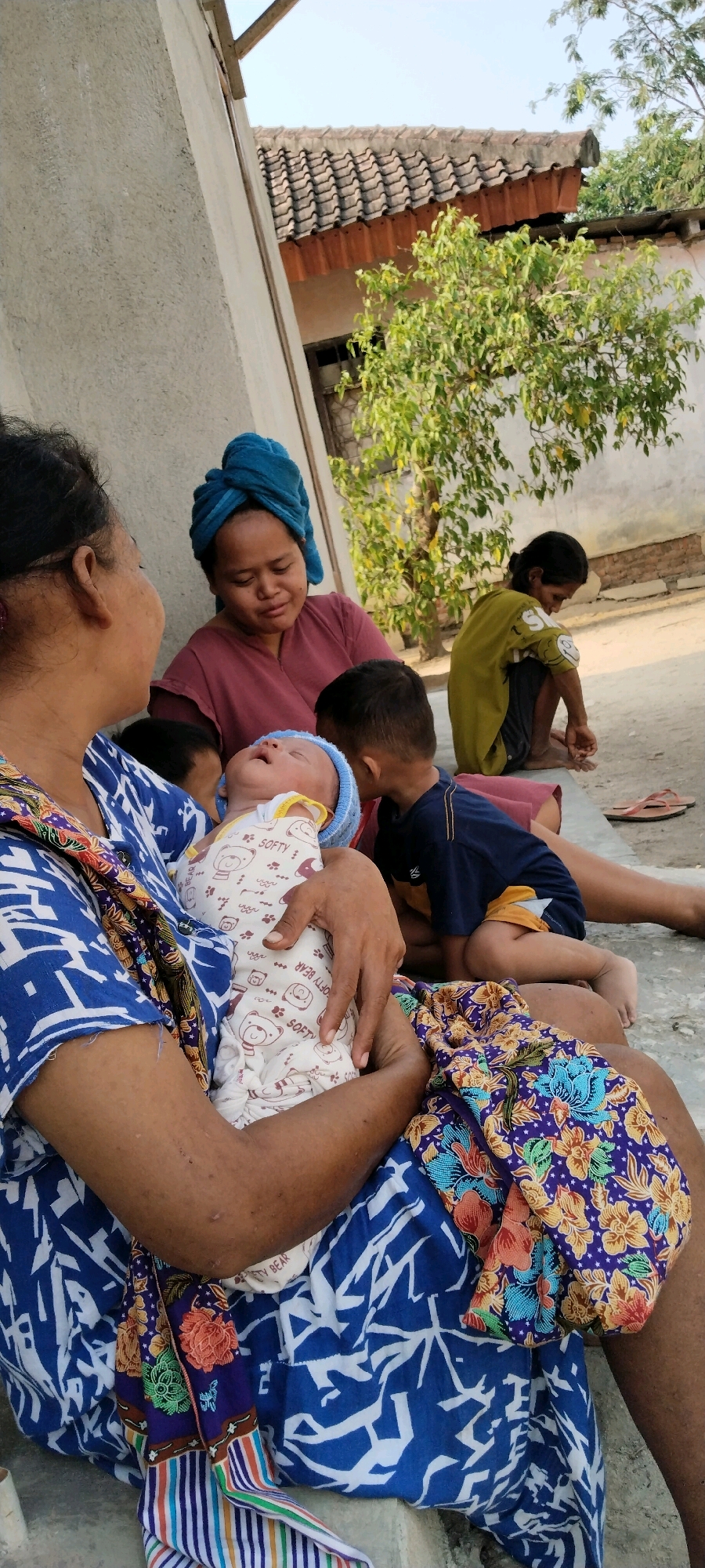 In an outdoor scene possibly in Tanjung Ratu Ilir, Indonesia, an older woman is holding a newborn baby swaddled in a white onesie with bear motifs. The baby is wearing a light blue hat. Beside her, a woman with a blue headscarf looks down at the baby. To the right, two young boys are present. One boy, wearing a dark blue shirt and yellow shorts, is looking towards the baby. Another child, wearing a green shirt, is sitting further back with their head bowed. The background shows a tiled roof and a tree, suggesting a rural or semi-urban setting. The ground appears to be concrete.