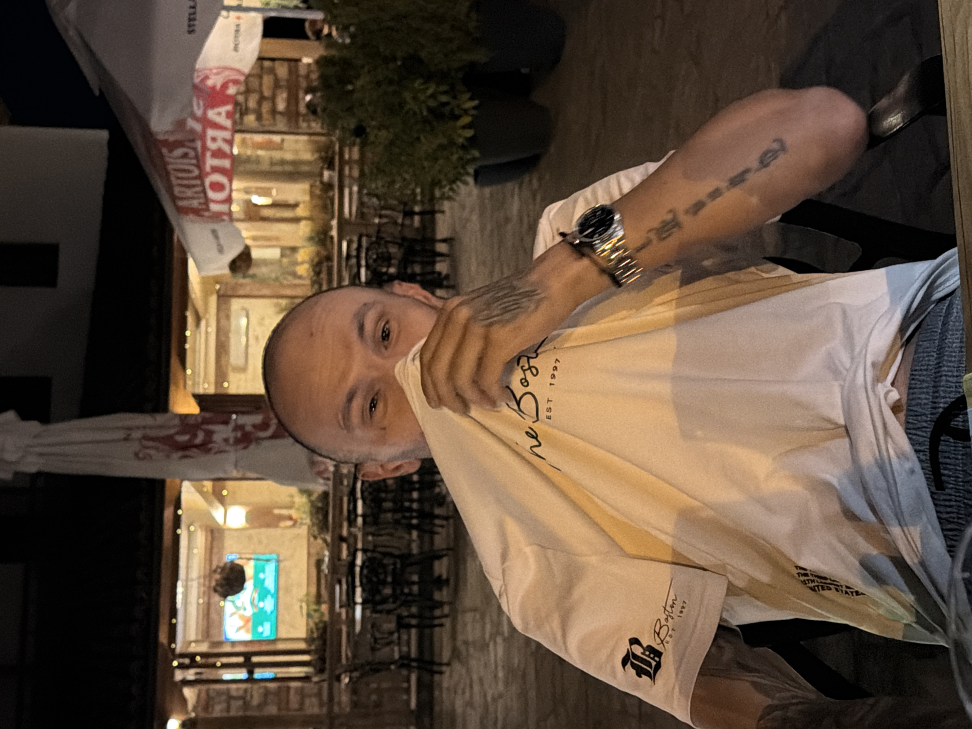 A man is seated at a table in an outdoor restaurant in Arbanassi, Bulgaria. He is wearing a white t-shirt and is partially obscuring his face with the fabric of the shirt. The man has a bald head and tattoos on his arm. He is wearing a watch on his left wrist. In the background, there are tables, chairs, and umbrellas, suggesting a dining area. The lighting indicates it is nighttime. There is signage visible in the background on an umbrella that reads "ARTOIS".