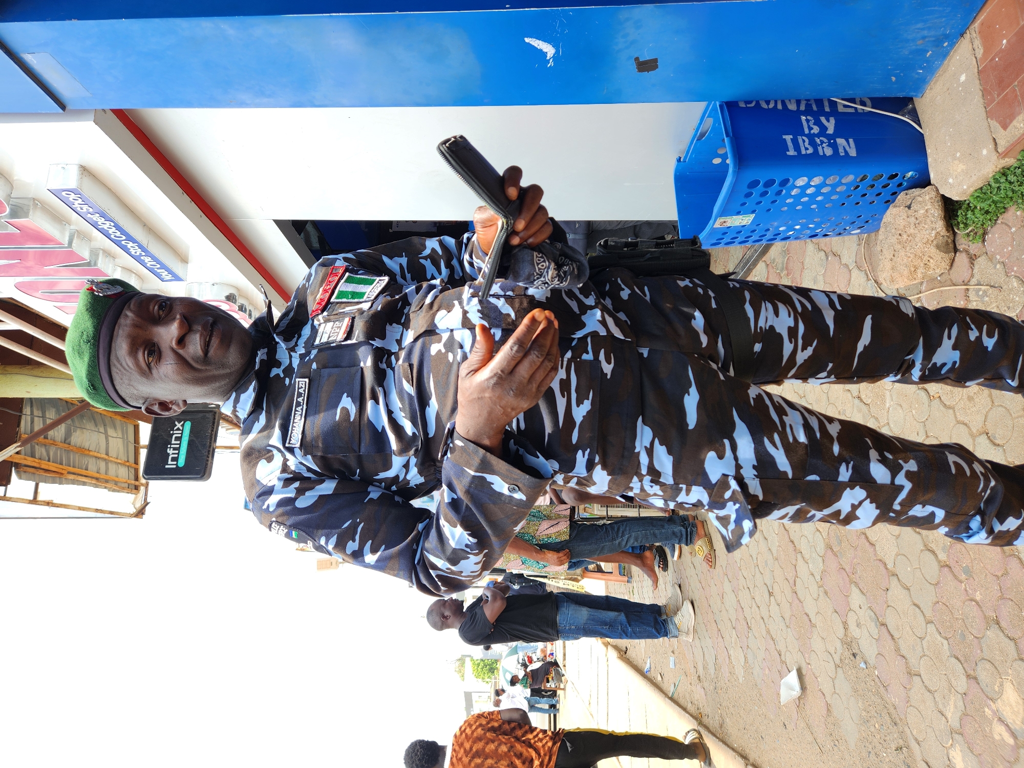The image shows a uniformed police officer standing on a patterned brick sidewalk, holding an open wallet in his hands. He is wearing a camouflage uniform with a green beret. His name tag reads "KOHANNA A.AZI". Behind him, there is a blue plastic basket with "DONATED BY IBN" printed on it. People are walking and standing in the background. An "Infinix" sign is visible on a nearby structure. The location is in a city. The specific details of the environment, such as buildings or street signs, are not clear enough to identify the precise location.