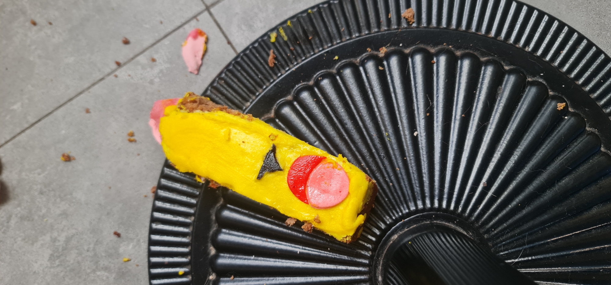 A partially eaten pastry resembling Pikachu lies on a black, ridged plastic plate. The pastry has a yellow frosting with a black triangle representing an eye and a red and pink circle for the cheek. The plate sits on a gray tiled surface. Debris from the pastry is scattered around the plate and on the tiled surface. No people are visible in the image. Đà Nẵng, Vietnam is not explicitly indicated but can be inferred from context (location data).