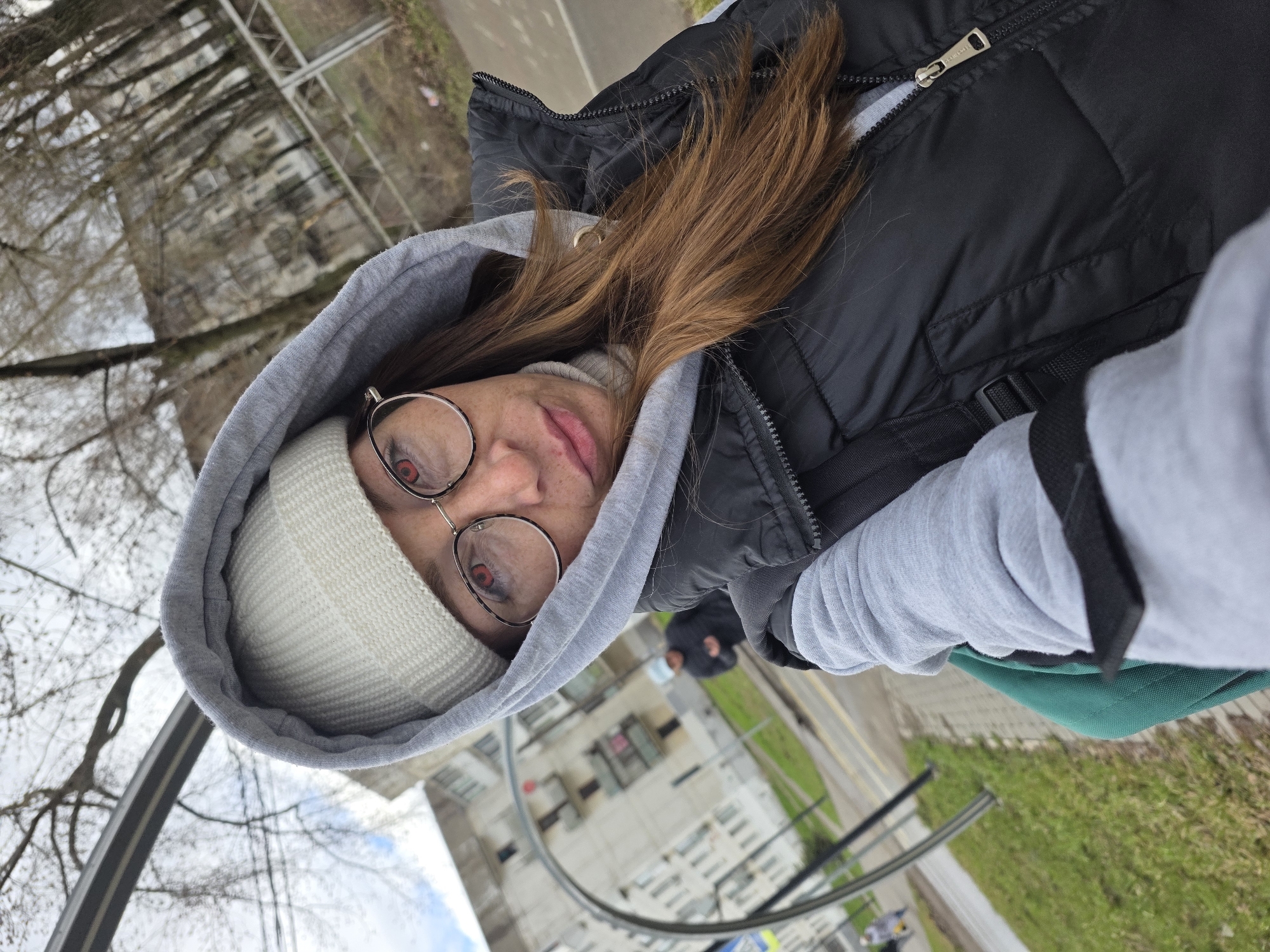 A close-up selfie, oriented diagonally, features a person looking towards the camera. The main subject, wearing a light gray hooded sweatshirt, a black puffer vest, and a light-colored knit beanie, has brown hair visible around the hood. They are wearing round, metallic-framed glasses, and a prominent red-eye effect is visible in their pupils. Their lips appear slightly chapped.
In the background, an overcast outdoor setting is visible, featuring bare trees, distant light-colored buildings, and a paved path or road. A metal railing, possibly a bicycle rack, is present in the midground. Another person, partially visible in the distance, wears a dark jacket and a light-colored face mask.
Notable details include a metallic zipper pull tab on the black puffer vest, which appears to have the letters "C&A" engraved on it. A black backpack strap and a piece of green fabric are also visible on the right side of the frame. The scene suggests a cool, daytime environment in an urban or park area.