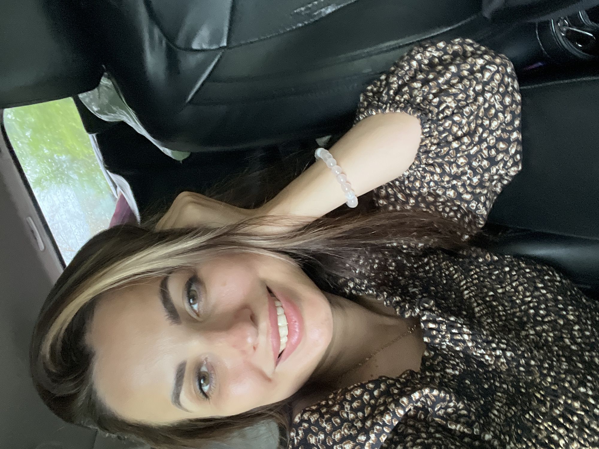 In an indoor, vehicle setting, a woman with brown hair and brown eyes is captured in a close-up, selfie-style photograph. She is wearing a dark, patterned top with a gold necklace. Her right arm is bent, with her hand resting near her face, and she is wearing a light-colored bracelet on her wrist. The background shows a black car seat and a window with greenery visible outside, suggesting it might be raining.