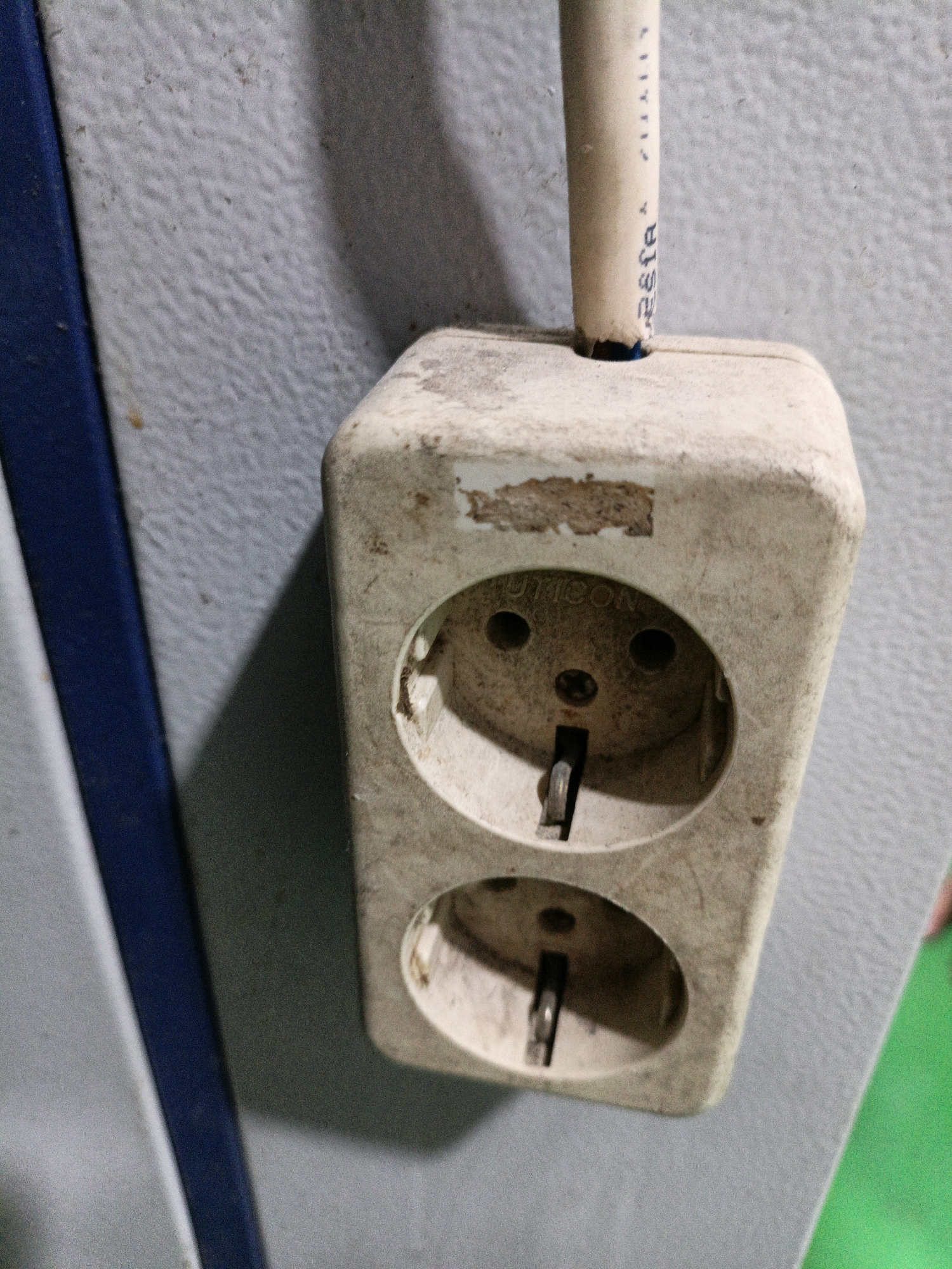 A close-up shot shows a dual electrical outlet that is dirty and appears to be old. The outlet is mounted on a textured, light gray wall. To the left of the outlet, a dark blue trim runs vertically along the edge of the wall. A white electrical cord extends from the top of the outlet and disappears upwards out of frame. There are some faint markings visible on the cord. The outlet itself has two circular receptacles, each with two openings for electrical prongs. The surface of the outlet is covered in a layer of dust and grime, with some discoloration and worn patches. The lighting in the scene creates a shadow on the wall to the right of the outlet. The image does not contain any people or specific identifiable landmarks that would indicate a precise location within North Jakarta, Indonesia.