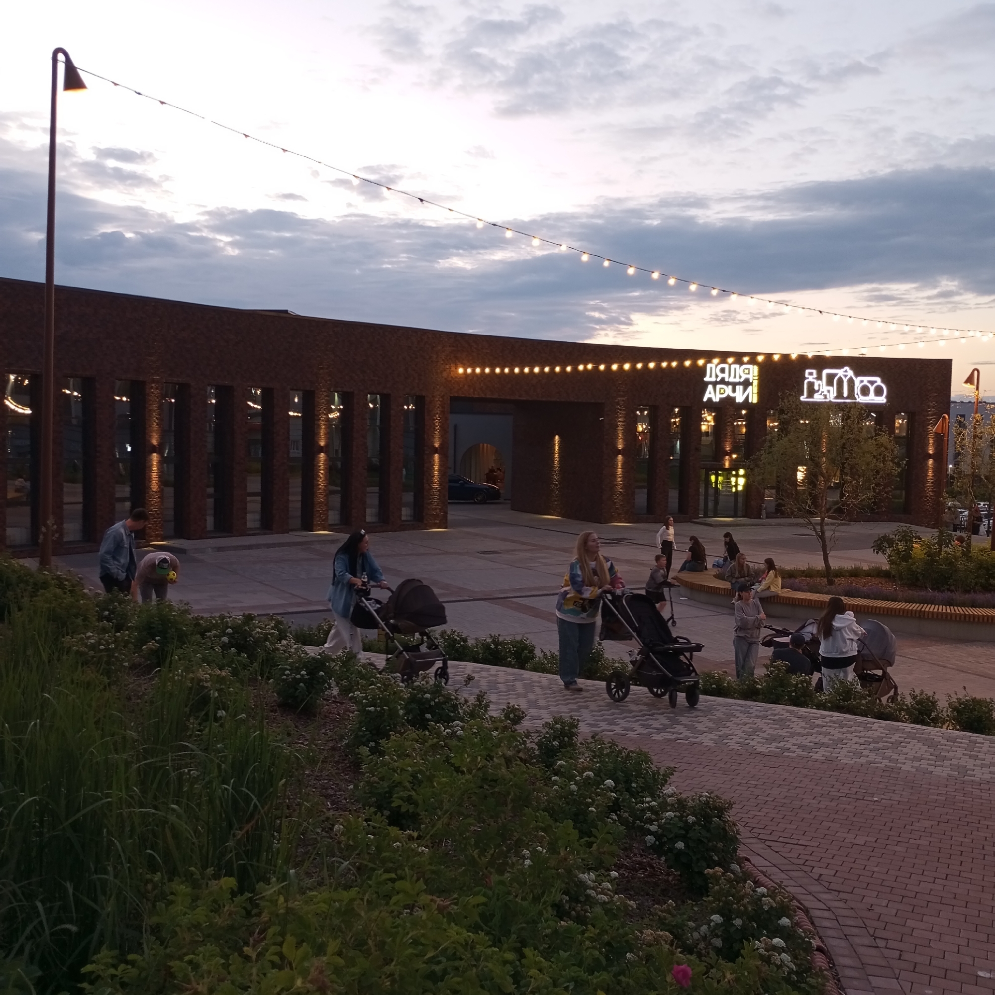 The image depicts an outdoor scene at dusk, likely in Izhevsk, Russia, given the signage. The primary structure is a modern, brick building with large, dark-tinted windows. Above the building's entrance, neon signage reads "Дядя Арчи" (Uncle Archie) and features an outline of various food items. String lights, some illuminated, are suspended across the sky, adding to the evening ambiance.
In the foreground and midground, several individuals are present, many with strollers. On the left, two men are near a grassy area; one is standing and looking towards the building, while the other is bending down, holding a yellow ball. Further to the right, a woman is pushing a stroller. In the center-right, another woman is also pushing a stroller. Behind them, a group of people are seated on a wooden bench, and a few more are standing or walking. A car is visible through an archway in the building. The ground is paved with a mosaic-like pattern of bricks. Lush green vegetation and flowering bushes line the pathway and the edge of the scene, suggesting a park or public plaza setting. The sky is cloudy, with hues of gray and hints of the setting sun.