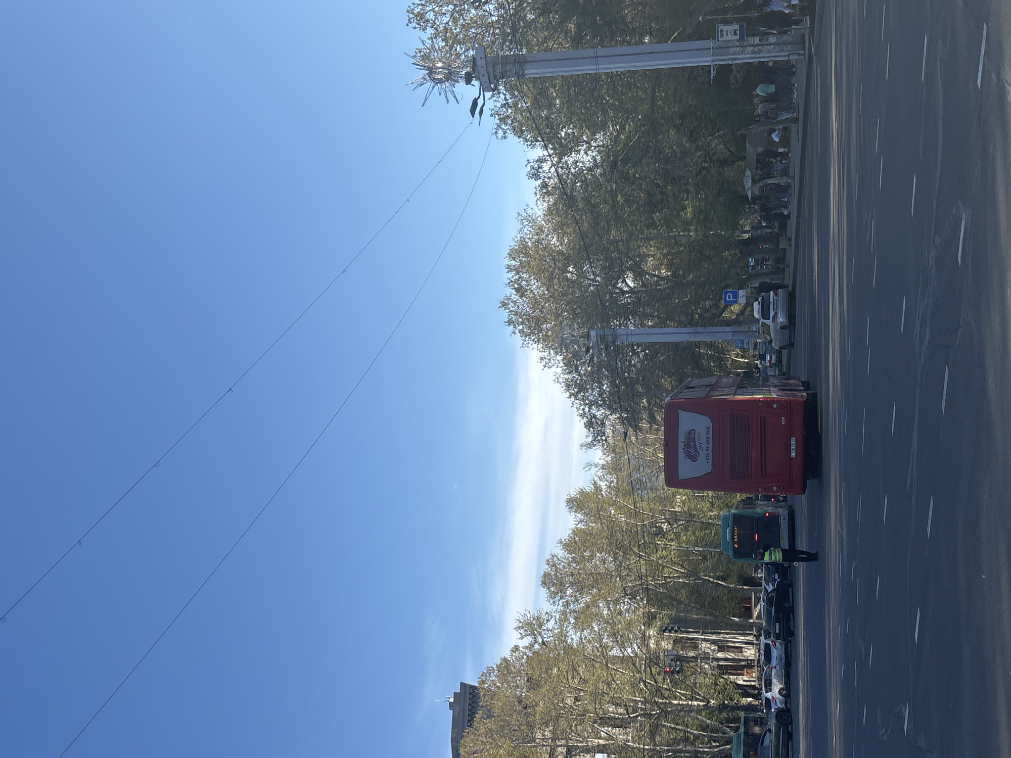 The image depicts a city street in Yerevan, Armenia, on a clear, sunny day. A red double-decker tour bus is partially visible, along with other cars and a green bus, all stopped in traffic. A traffic officer in a yellow vest is standing near the vehicles. Trees line the street, and utility poles with decorative lighting are present. Street signs and parking indicators can be seen. In the distance, a building can be observed. The sky is clear blue with some faint clouds.
