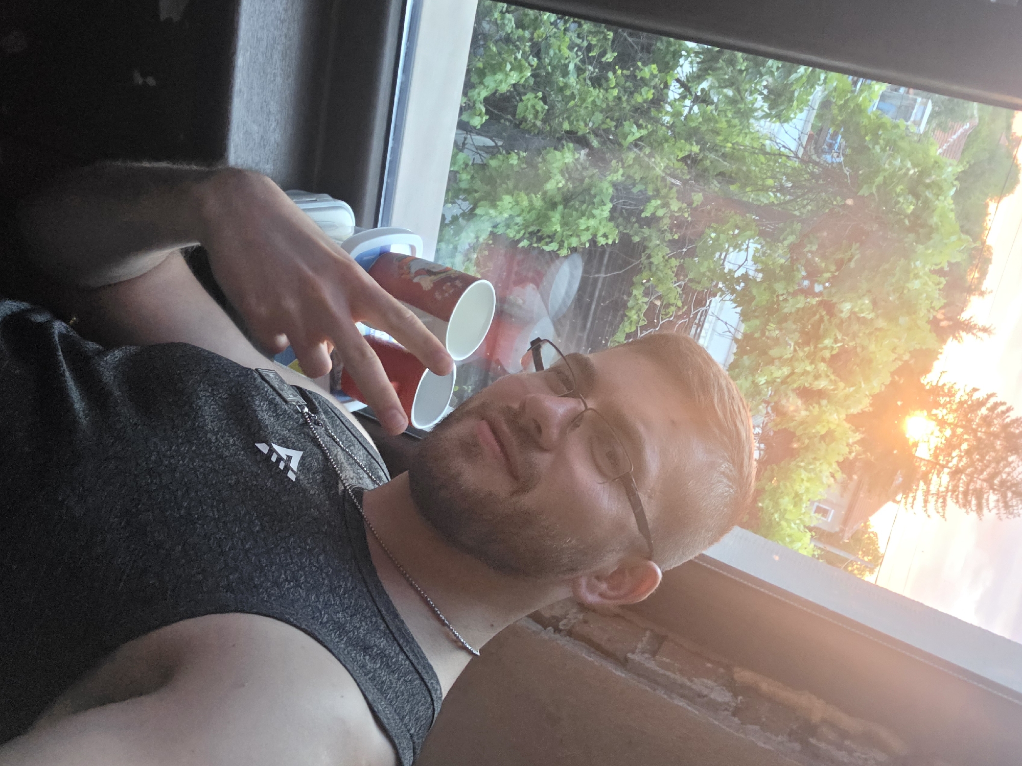 A man with fair skin, short reddish-blonde hair, and a beard is indoors near a window. He is wearing glasses and a dark-colored tank top with a small logo on the chest. A thin silver chain is visible around his neck. The man is holding up a peace sign with his left hand near two paper cups sitting on a window sill.
Through the window, there is a view of green trees, houses, and a sunset with an orange glow. The location is difficult to pinpoint exactly, but appears to be a residential area. There is nothing in the image indicating that the location is Yagodovo, Bulgaria.