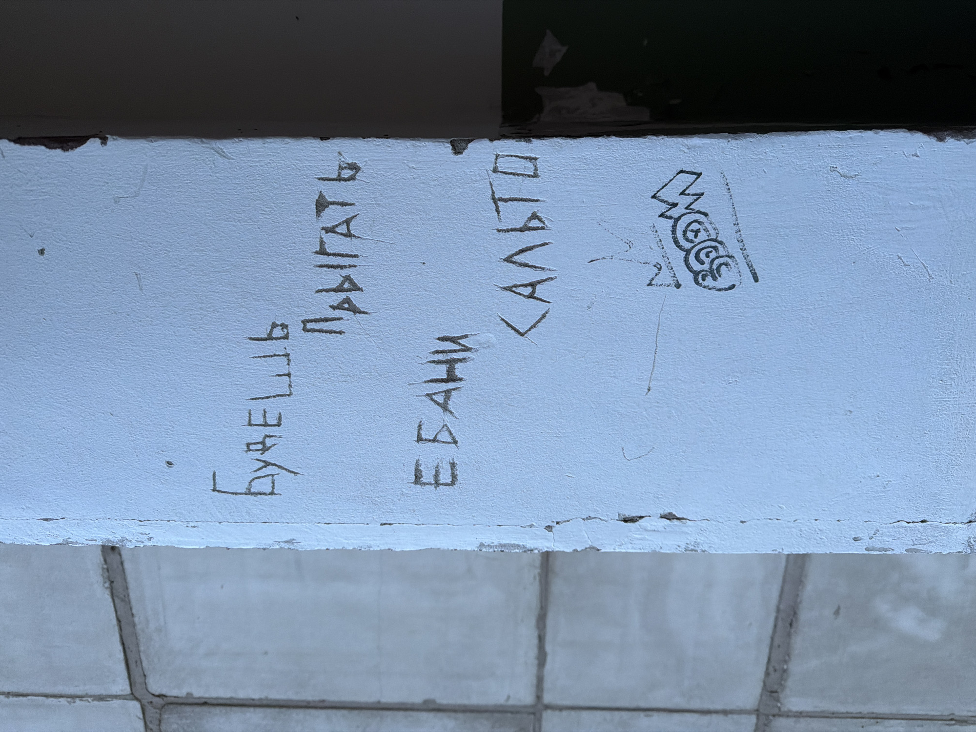 The image displays a light blue or off-white painted concrete pillar or wall segment located in Chaikovsky, Russia. The painted surface exhibits wear, chipping, and vertical cracks. Engraved or scratched into the paint are two lines of Cyrillic text. The upper line reads "БУДЕШЬ ПРЫГАТЬ", positioned slightly left of center. Below it, to the right, the second line reads "ЕБАНИ САЛЬТО". Below this text, a black marker graffiti tag is present, featuring a stylized "BOOM" with a crown symbol directly above it. To the left of the pillar, a light grey tiled wall with dark grout lines is visible. The right edge of the pillar transitions into a dark, shadowed background, where a section of a dark green or brown wall is discernible at the bottom. No individuals are present in the scene.