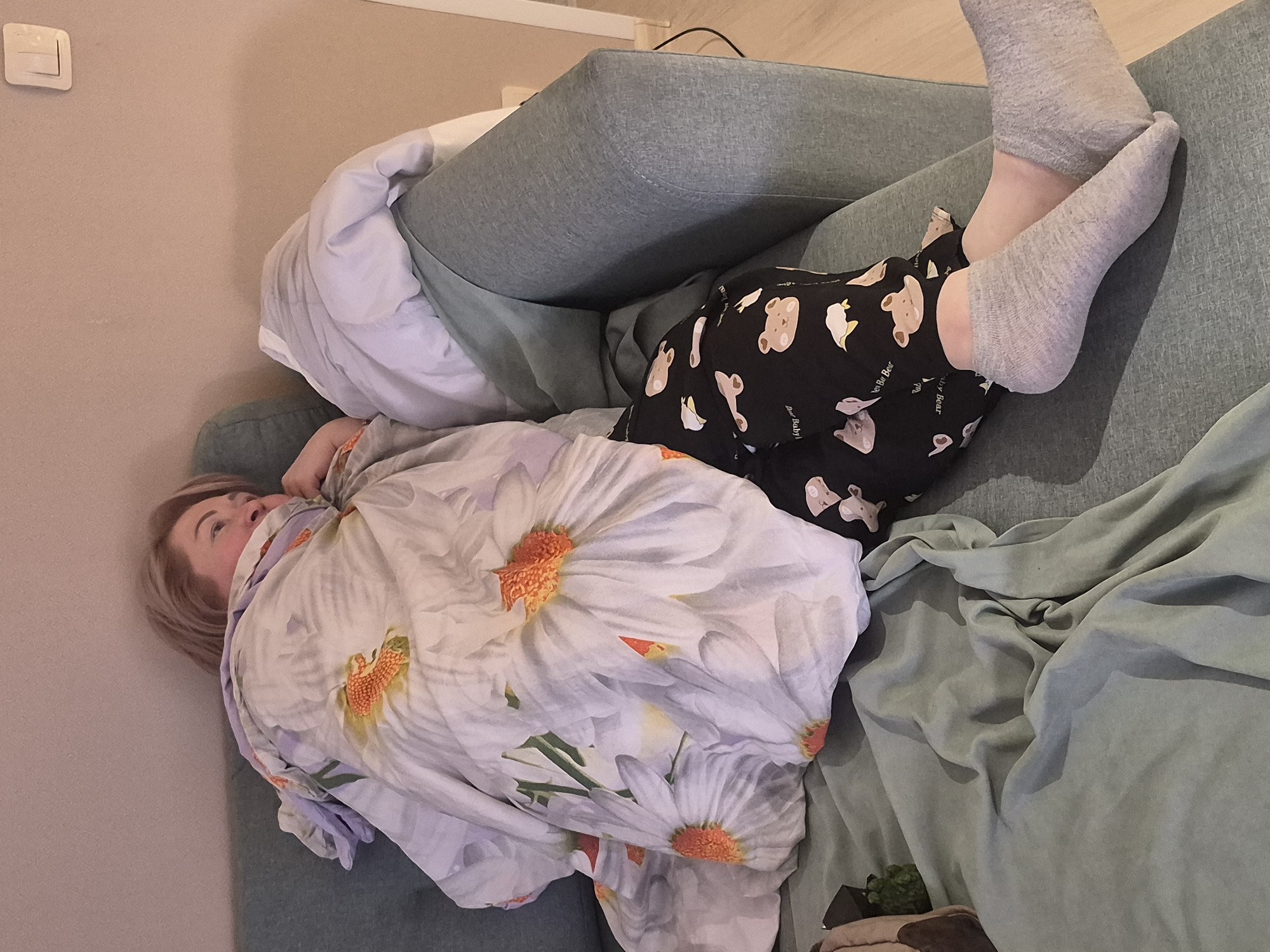 A person lies on a light green couch, covered with a floral-patterned blanket. The person wears gray socks and black pajamas with a white bear design. Part of a white pillow and a light gray cushion are visible on the couch. The photo appears to be taken indoors, possibly in a residential setting. No specific details link the image to Chaikovsky, Russia.