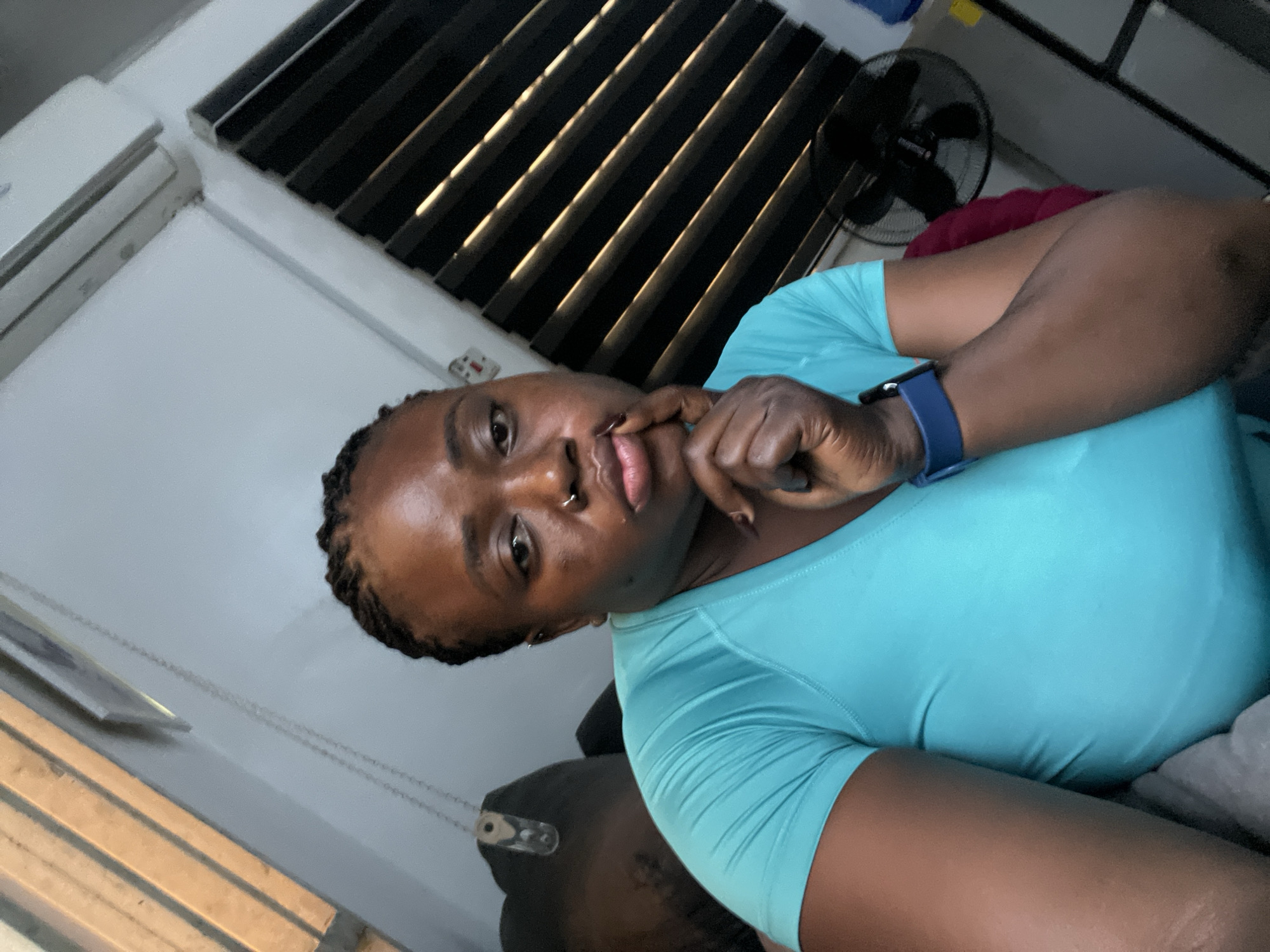 The image shows a close-up of a Black woman in a turquoise t-shirt, with her head tilted and her hand near her mouth. She has dark braided hair, a nose ring, and is wearing a blue smartwatch on her left wrist. The background is dimly lit and appears to be an indoor setting. Behind her, there are horizontal blinds which are partially illuminated, suggesting a light source outside or from a window. To the left of the blinds, a white air conditioning unit is visible. A black fan is also visible in the background, to the right of the blinds. The overall lighting and ambiance suggest it might be evening or a dimly lit room. The woman's expression is neutral or thoughtful. There are no visible text elements in the image. The context provided indicates the location is Abuja, Nigeria.