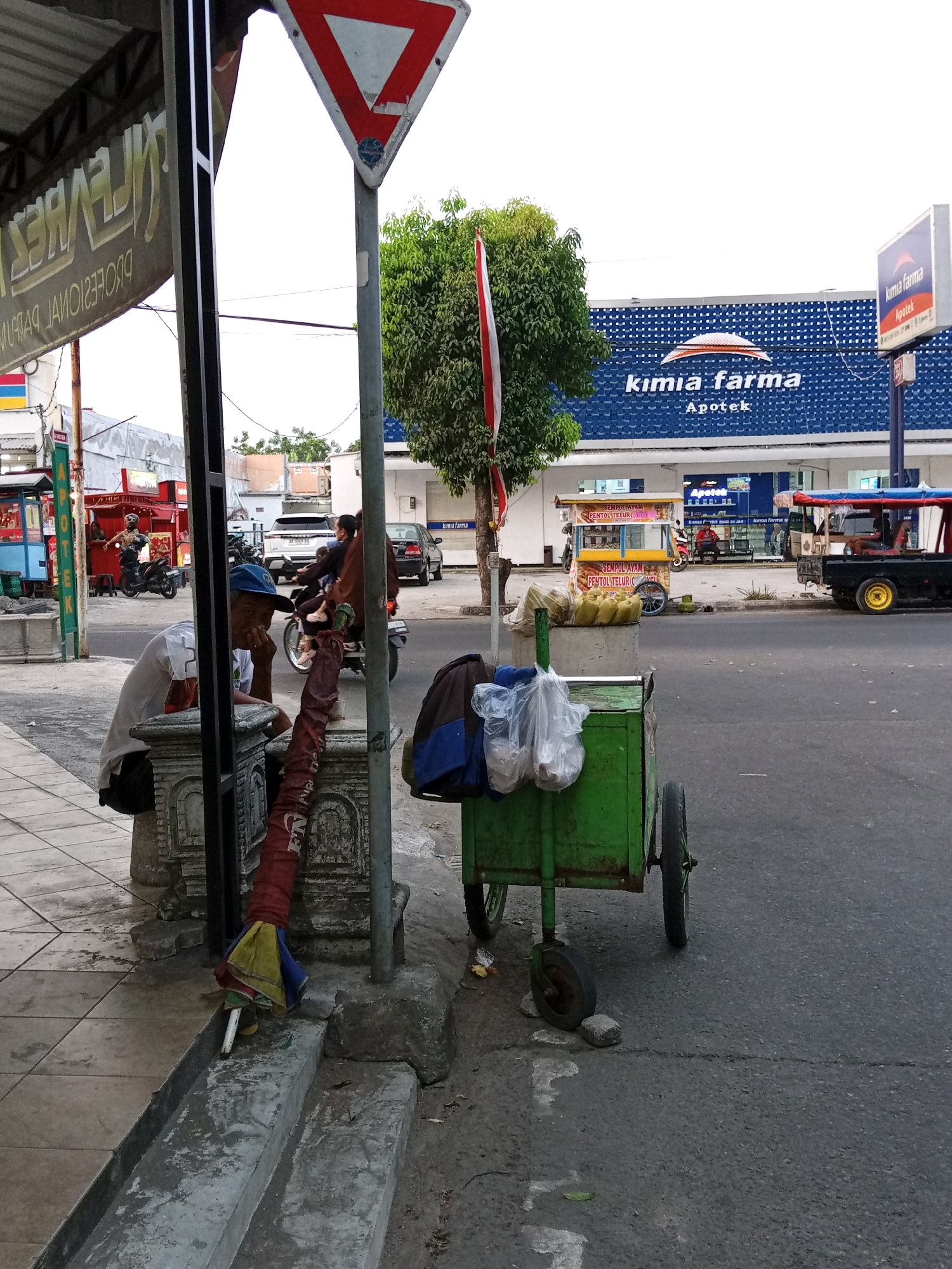 This is a street scene in Sukoharjo, Indonesia, near a Kimia Farma pharmacy. In the foreground, a man wearing a blue cap and a white t-shirt sits on a concrete structure, looking to his left. Beside him is a collapsed umbrella with red, yellow, and blue sections. To the right of the man, a green wheeled cart is parked on the asphalt. This cart holds several plastic bags containing items, and on its side are two dark blue and brown bags. In the background, the blue building of Kimia Farma is visible with its white signage. A red and white Indonesian flag flies from a tree in front of the building. Other street elements include a give way sign, a motorbike with a rider, a food cart, and various vehicles on the road. The street surface is asphalt, and the sidewalk is made of concrete tiles and stone.