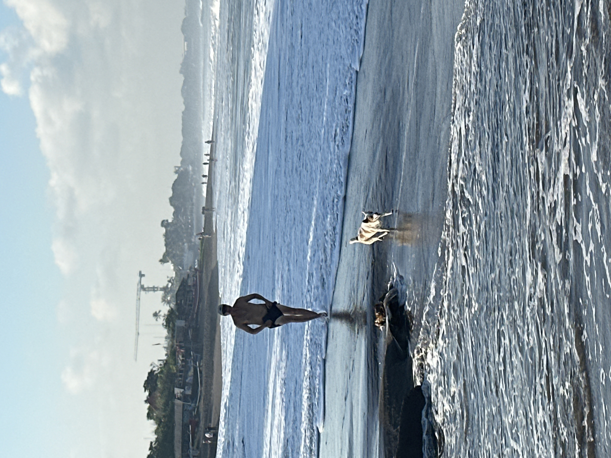 A man in dark shorts and a dark cap is walking towards the camera on a wet beach, with waves breaking around his feet. Beside him and slightly ahead, a small, light-colored dog with dark markings runs. In the background, a long line of people walk along the shore, with a hazy coastline and a construction crane visible in the distance. The sky is bright with scattered clouds. The scene is captured on a beach in North Kuta, Indonesia.