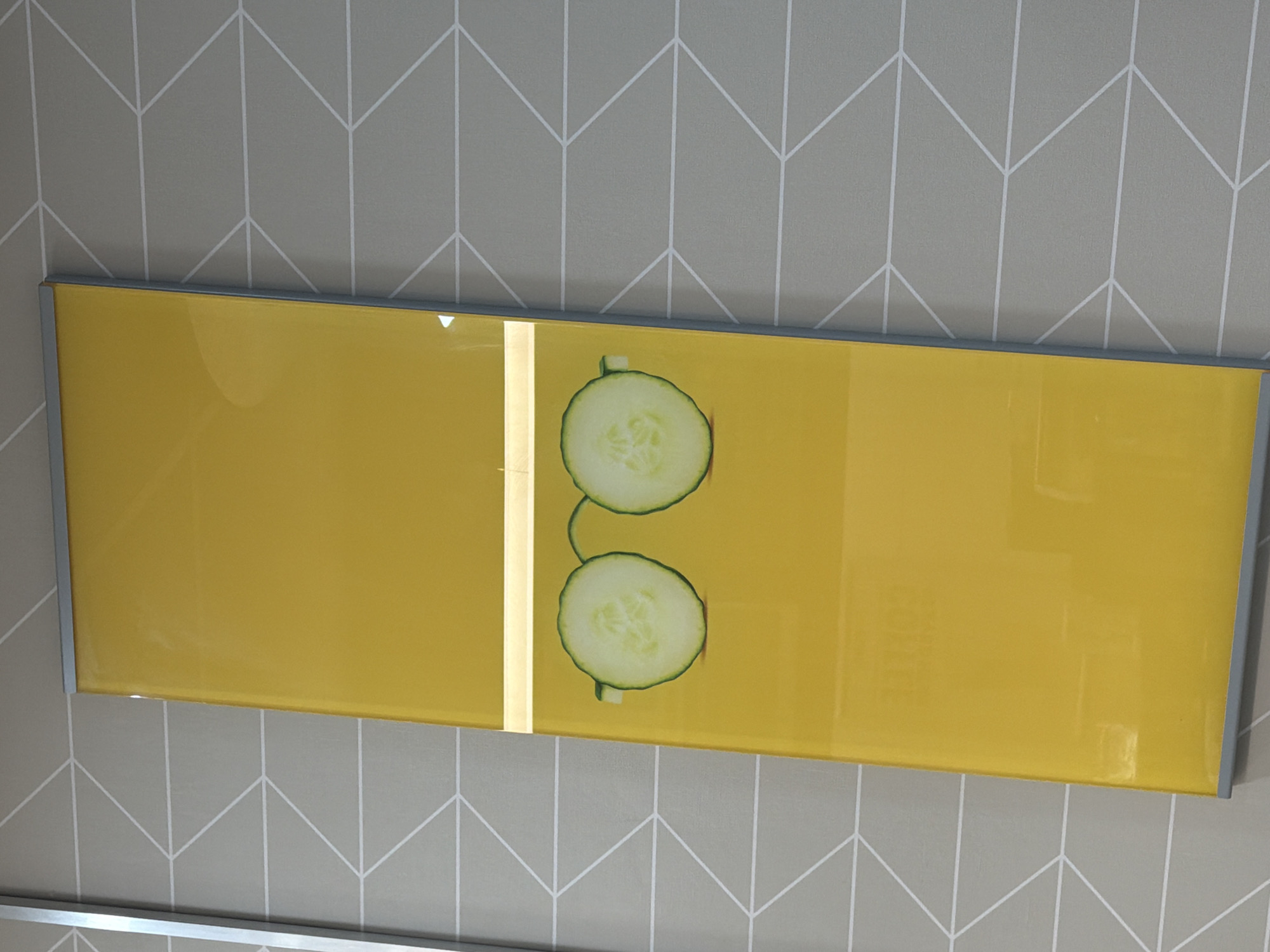 A rectangular picture with a silver frame hangs on a wall with a gray geometric pattern. The picture's background is yellow and divided into two sections by a vertical gold stripe. In the right section of the picture, two circular cucumber slices are stacked vertically. The picture is mounted on the wall in an indoor environment, likely a commercial establishment. There is no information about the location being in Pity Me, United Kingdom.