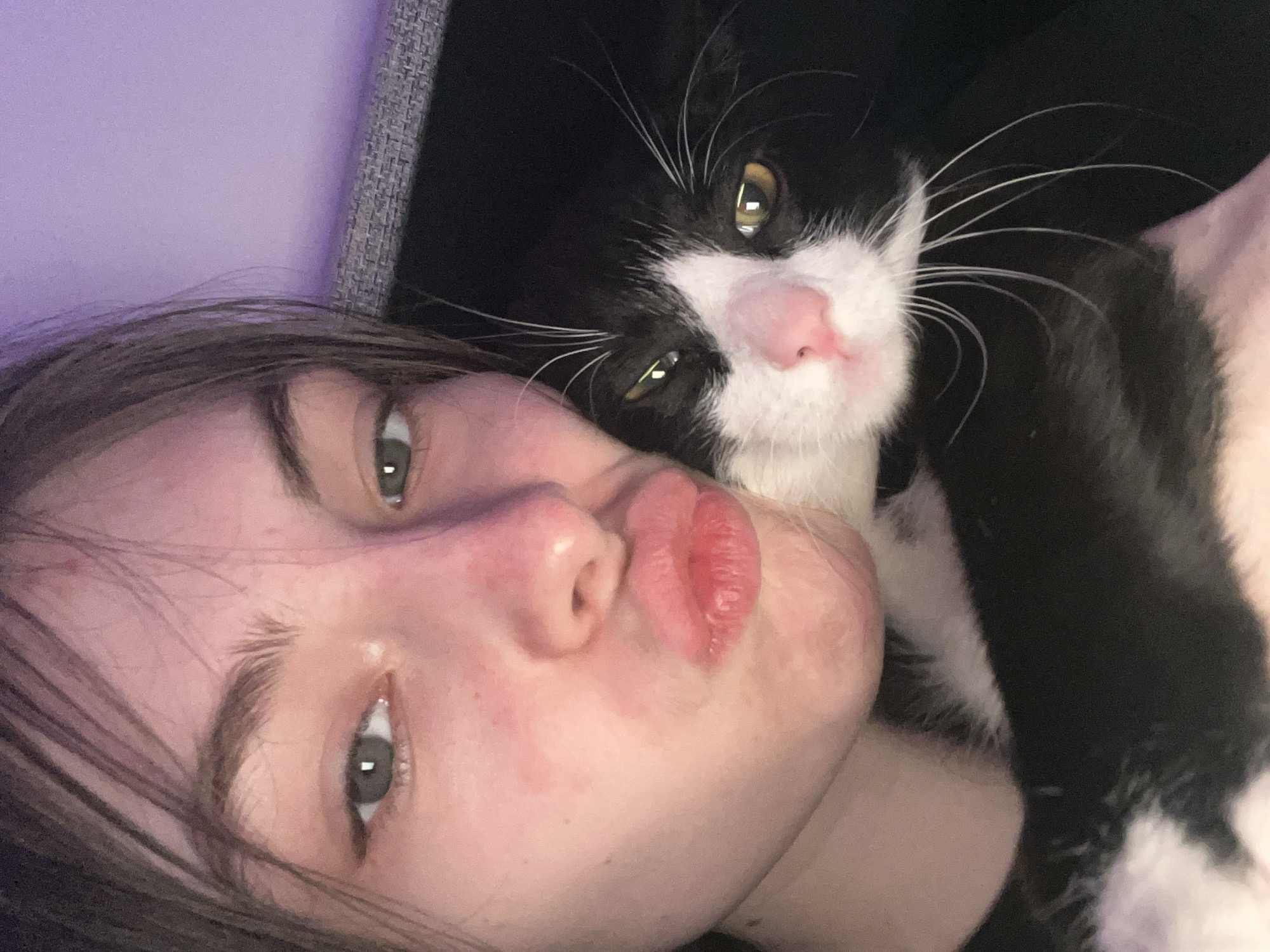 A young woman with light skin, green eyes, and brown hair is pictured in a close-up selfie, pursing her lips. Adjacent to her face is a black and white cat, its face also prominent in the frame. The cat has yellow eyes and white whiskers. The background is indistinct, showing shades of purple and gray. The image does not depict Saint Petersburg, Russia.