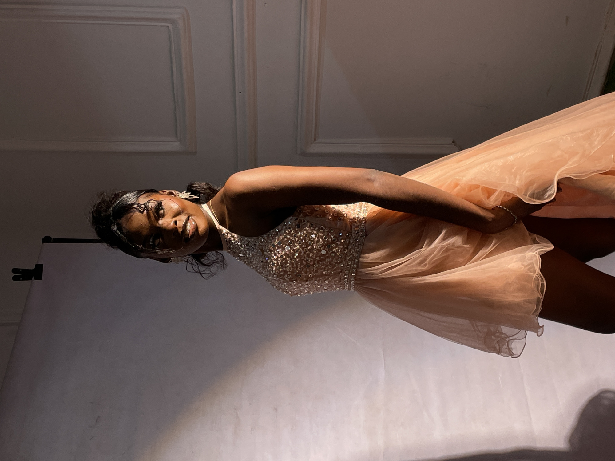 The original image, which is rotated 90 degrees counter-clockwise, depicts a young Black woman in what appears to be an indoor studio or event setting in Barakin Akawo, Nigeria. She is the sole subject, standing and looking directly at the camera with a confident, slight smile.
The woman is wearing an elegant, light peach or nude-colored party dress. The bodice of the dress is intricately embellished with numerous shimmering sequins or beads, creating a sparkling effect. The short skirt is voluminous and layered, made from a sheer, flowing fabric like tulle or organza, which gives it an airy appearance. She accessorizes with delicate, ornate earrings and a subtle bracelet on her wrist. Her hair is styled and pulled back, and she has visible makeup, including eyeliner and lip gloss, enhancing her features.
The background consists of plain walls with some architectural molding or wainscoting visible, suggesting a room with classic design elements. To the right of the subject (left in the rotated image), a soft, lighter backdrop or a large white surface with a stand and clip is visible, indicating a controlled photographic environment. The lighting is focused, casting highlights on her skin and the dress, particularly on the embellished bodice, and creating some soft shadows in the background. The overall impression is that of a posed portrait, possibly for a special occasion or fashion shoot. No visible text is present in the image.