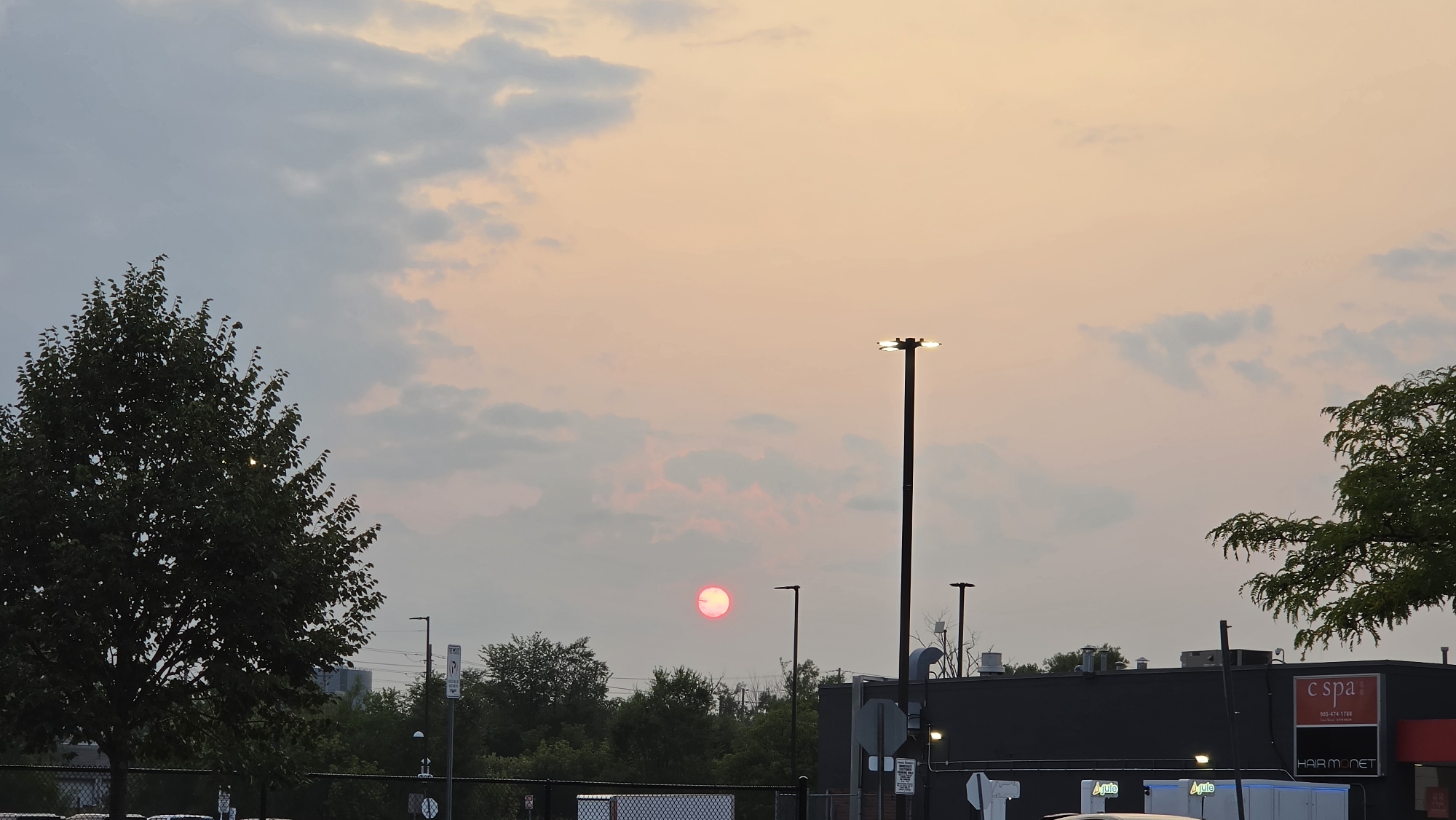 The image depicts a dusk or dawn scene in the city of Markham, Canada. The sky is filled with a gradient of colors, transitioning from a pale blue at the top to soft peachy and pink hues towards the horizon, with scattered clouds present. A bright, reddish sun is visible in the sky, partially obscured by a faint haze or clouds, giving it a diffused appearance.

In the foreground, the silhouette of a large, leafy tree is visible on the left side of the frame. On the right, a dark-colored commercial building with a red sign that reads "c spa" and "HAIR MONET" is partially visible. Several streetlights are also present, one of which is tall and prominent with multiple lamps. A chain-link fence runs along the bottom of the image, with what appear to be parked vehicles behind it. Green trees and vegetation form a line behind the fence and in front of the building. The overall environment suggests an urban or suburban setting at either sunrise or sunset.