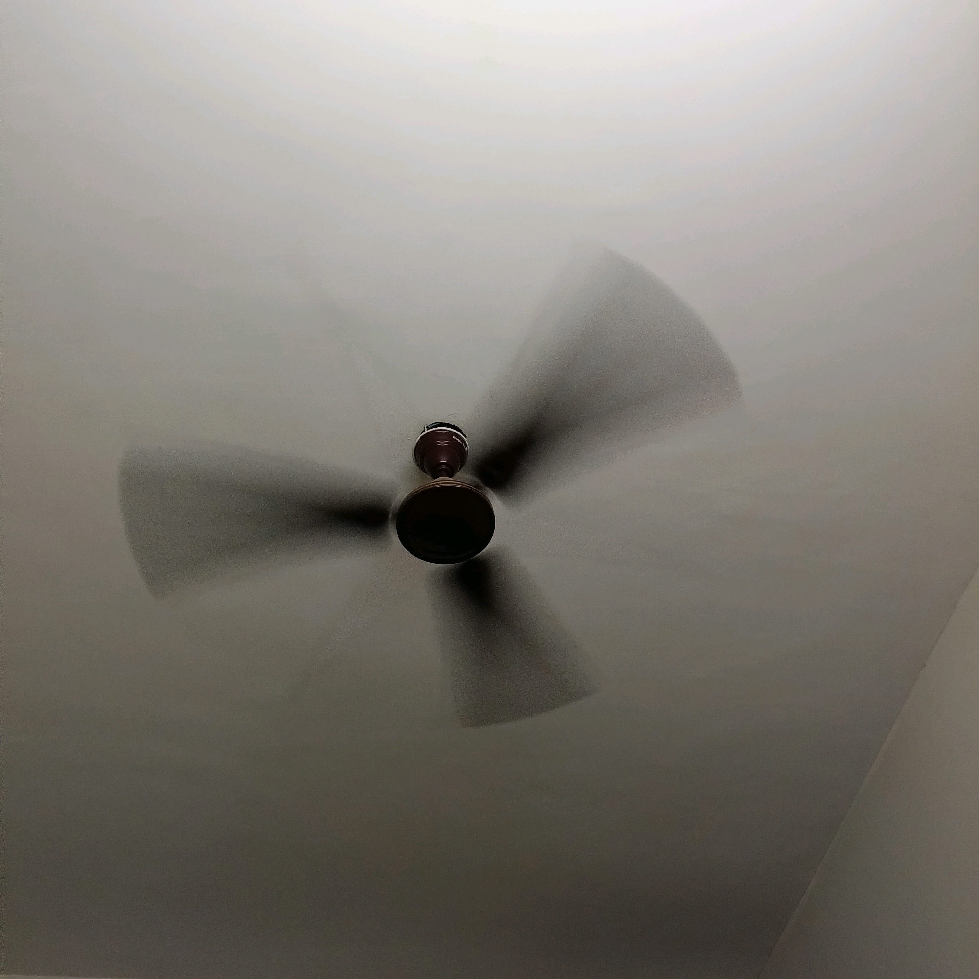 The image shows a ceiling fan in motion, mounted on a white ceiling. The fan has a dark brown motor housing and four blades that are blurred due to the motion. The lighting is dim, creating shadows on the ceiling. There are no people or other discernible objects in the image. The scene is indoors, and the time of day is indeterminate due to the artificial lighting and lack of natural light sources. The location in Bengaluru, India cannot be determined from the image alone.