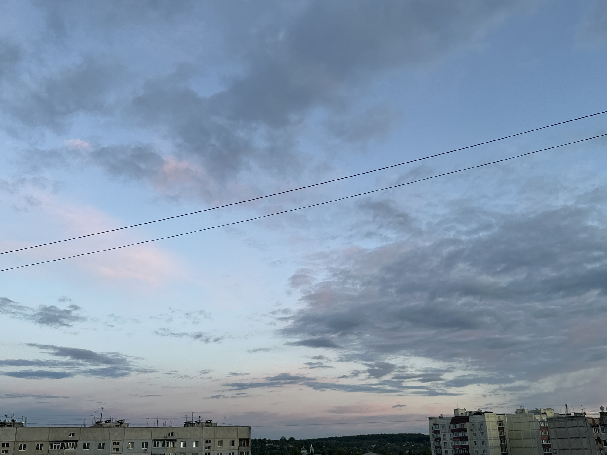 The image depicts a cityscape at dusk or dawn. Two multi-story residential buildings frame the bottom of the image. The foreground shows a backdrop of green trees, possibly a park or residential area. Overhead, the sky is filled with a mix of light blue and gray clouds, with a hint of pink hues around the edges, suggesting the presence of sunlight. A pair of power lines cross the sky horizontally in the upper part of the frame. The location is in the city of Unknown, Unknown.