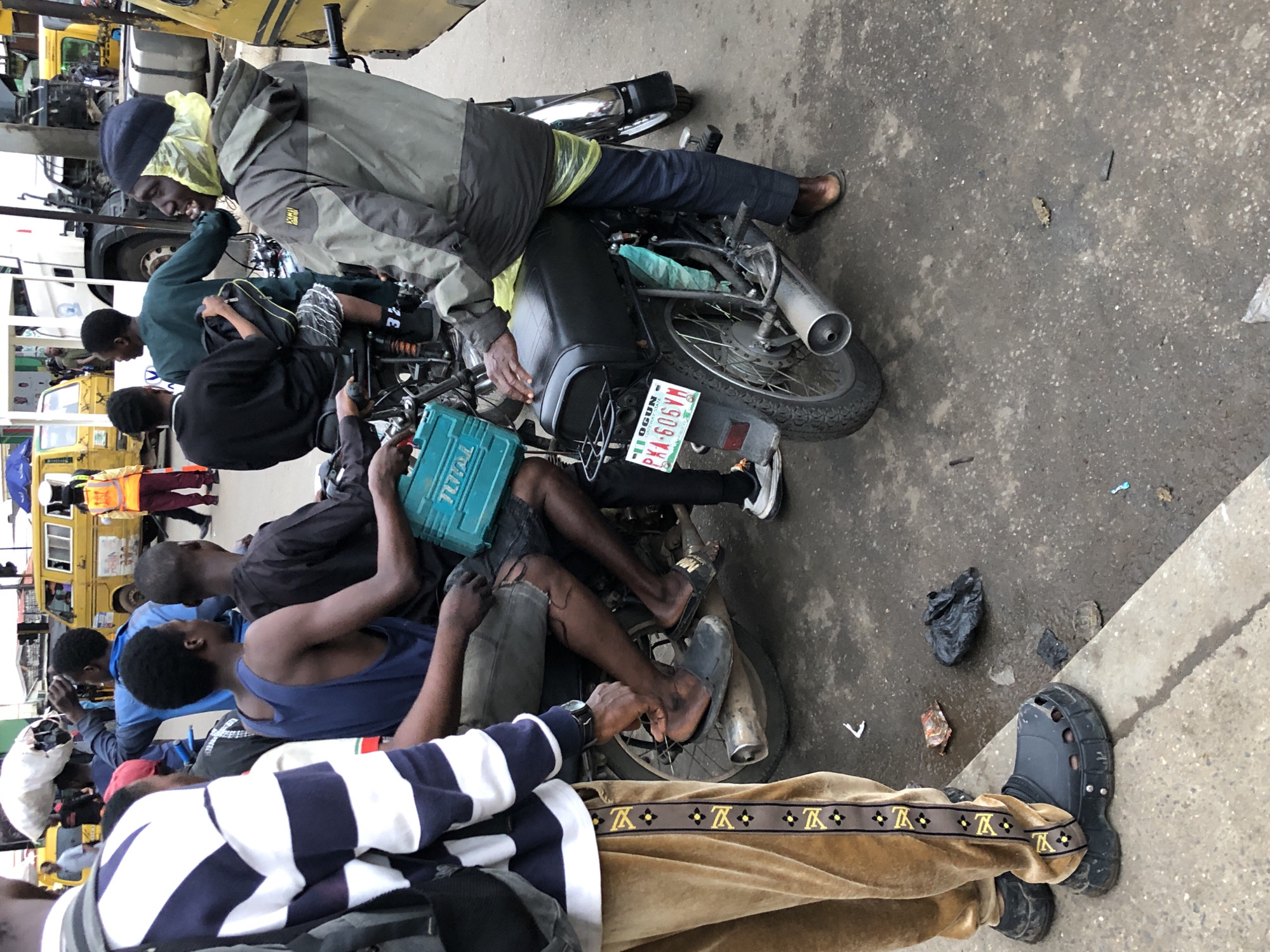 This image captures a bustling street scene in Lagos, Nigeria, likely in a busy commercial or transport hub. The foreground features a person wearing distinctive tan track pants with a gold pattern and oversized black crocs, standing next to a parked motorcycle. The motorcycle has a turquoise toolbox secured to its rear and a license plate reading "OGUN PKA 606VM".
Several other individuals are visible around the motorcycle, suggesting a common mode of transport or a gathering point. One man, wearing a grey jacket and a yellow plastic bag covering his head, sits on the motorcycle, facing towards the right of the frame. Another man in a dark blue tank top and shorts is seated behind him. Behind them, other individuals are either seated or standing, partially obscured. The background reveals other vehicles, including a yellow minibus, and a white truck, indicative of active traffic and commerce. The ground is paved and appears wet in places, with scattered debris, suggesting a recent rain or general street conditions. The overall impression is one of everyday life in a densely populated urban environment.