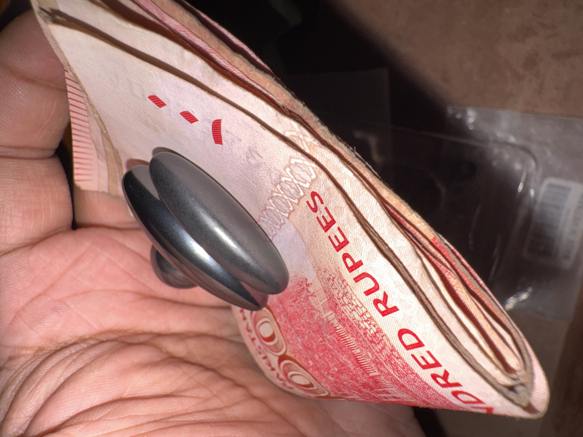 A hand holds a folded stack of Pakistani Rupees secured with a metallic money clip. The currency is predominantly pink and red in color. The background is dark and blurry, but it appears to be an indoor setting. The location is unknown.