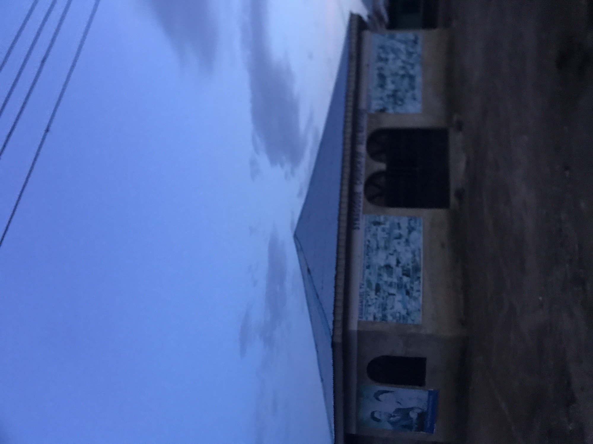 The image depicts a single-story building at dusk or dawn in Rafin Par, Nigeria. The structure features light-brown colored walls and a light-blue corrugated metal gable roof. The facade includes two dark, arched entrances or windows at its center. On the leftmost wall, a large poster displays two individuals, one male in a suit and one female. Above the central section of the building, partially visible text reads "EMMANUEL TV" and "SYNAGOGUE CHURCH OF ALL NATIONS." Below this text, and extending across portions of the front wall, are collages composed of numerous small, light-toned images set against a blue background. The immediate foreground consists of dark, unpaved ground. The upper half of the scene is dominated by a deep blue sky with scattered, faint gray clouds. Several dark overhead power lines traverse the upper right quadrant of the sky.