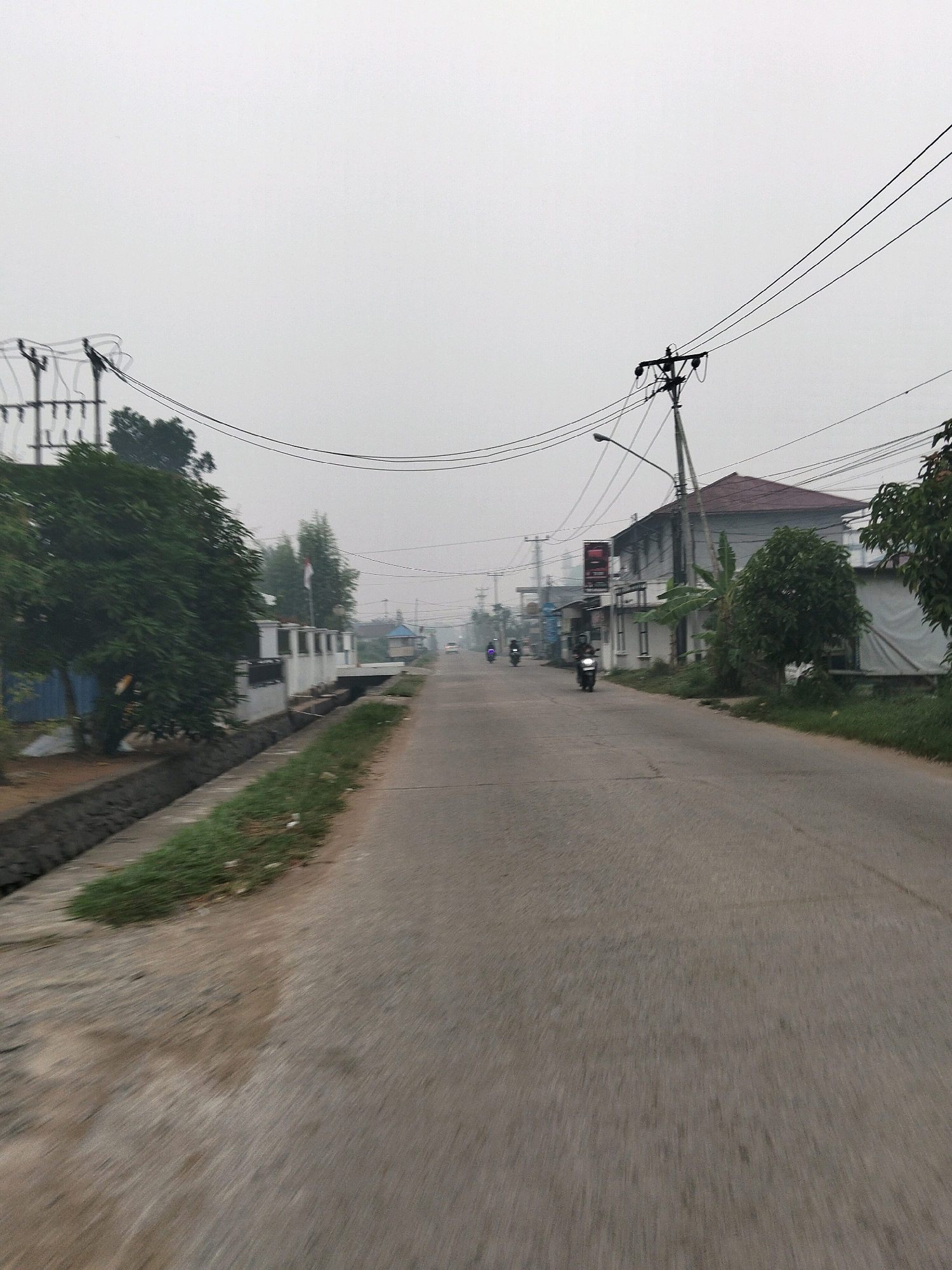 The image captures a street scene in Arang Limbung, Indonesia, under an overcast sky, likely early morning or during misty conditions. The foreground is dominated by a paved road with blurred motion, suggesting the photograph was taken from a moving vehicle. To the left, a concrete drainage ditch runs alongside the road, with some patchy grass growing within it and on the embankment. Beyond the ditch, a large, dense green tree is visible, with a section of a blue structure behind it. Further down the road on the left, a white-walled building with a series of pillars and a connecting bridge is seen, with a flagpole displaying the Indonesian flag.
On the right side of the road, a two-story building with a reddish-brown roof stands, partially obscured by vegetation. In front of this building, a large tree with dark green foliage grows. Further down the street on the right, the facades of several other buildings are visible, including what appears to be a shop or small business with signs.
Several individuals on motorcycles are present. One motorcycle with a rider is prominently featured in the middle ground, traveling away from the camera. Two other motorcycles, one with a rider, are visible further down the road on the left, also moving away from the camera. Utility poles with electrical wires crisscross the sky above the scene. The overall atmosphere is hazy and subdued, typical of a diffused light condition.