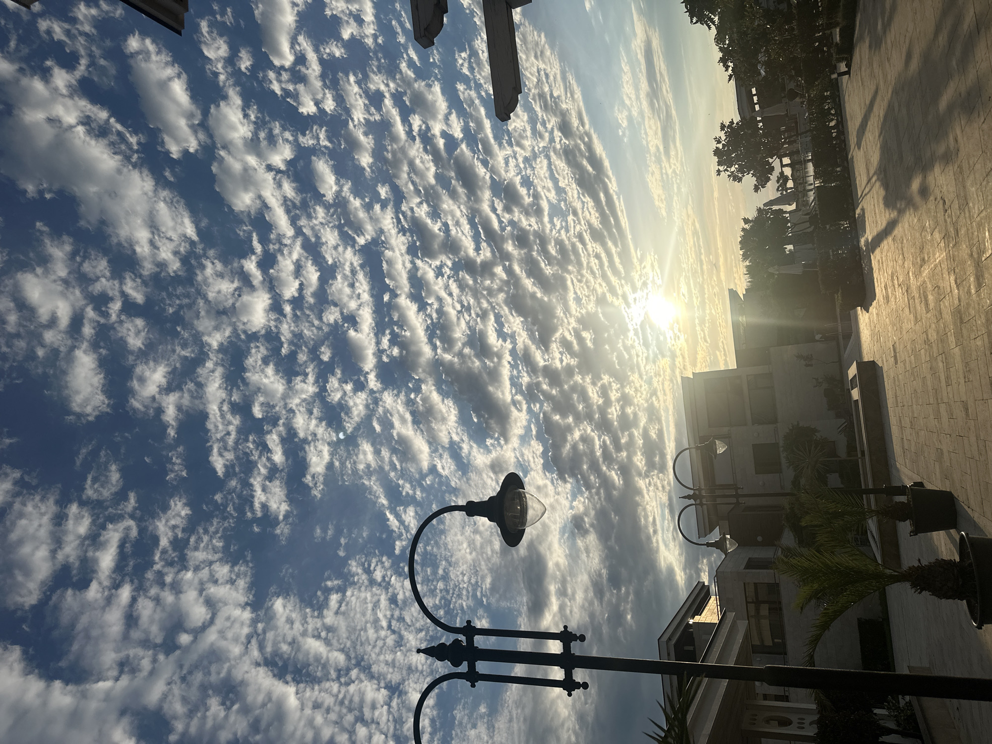 The image shows a picturesque view of the sky over Sozopol, Bulgaria. The sky is filled with a tapestry of white, fluffy clouds against a vibrant blue backdrop. The sun is low in the sky, casting a warm, golden glow and creating long shadows on the paved ground below. In the foreground, a classic black lamppost with a glass lantern stands tall, silhouetted against the illuminated clouds.
Further back, modern architectural structures with light-colored facades are visible, suggesting a developed area, possibly a resort or residential complex. Palm trees in pots add a touch of greenery and tropical flair to the scene. The overall ambiance is serene and tranquil, capturing the beauty of a late afternoon or early morning sky.