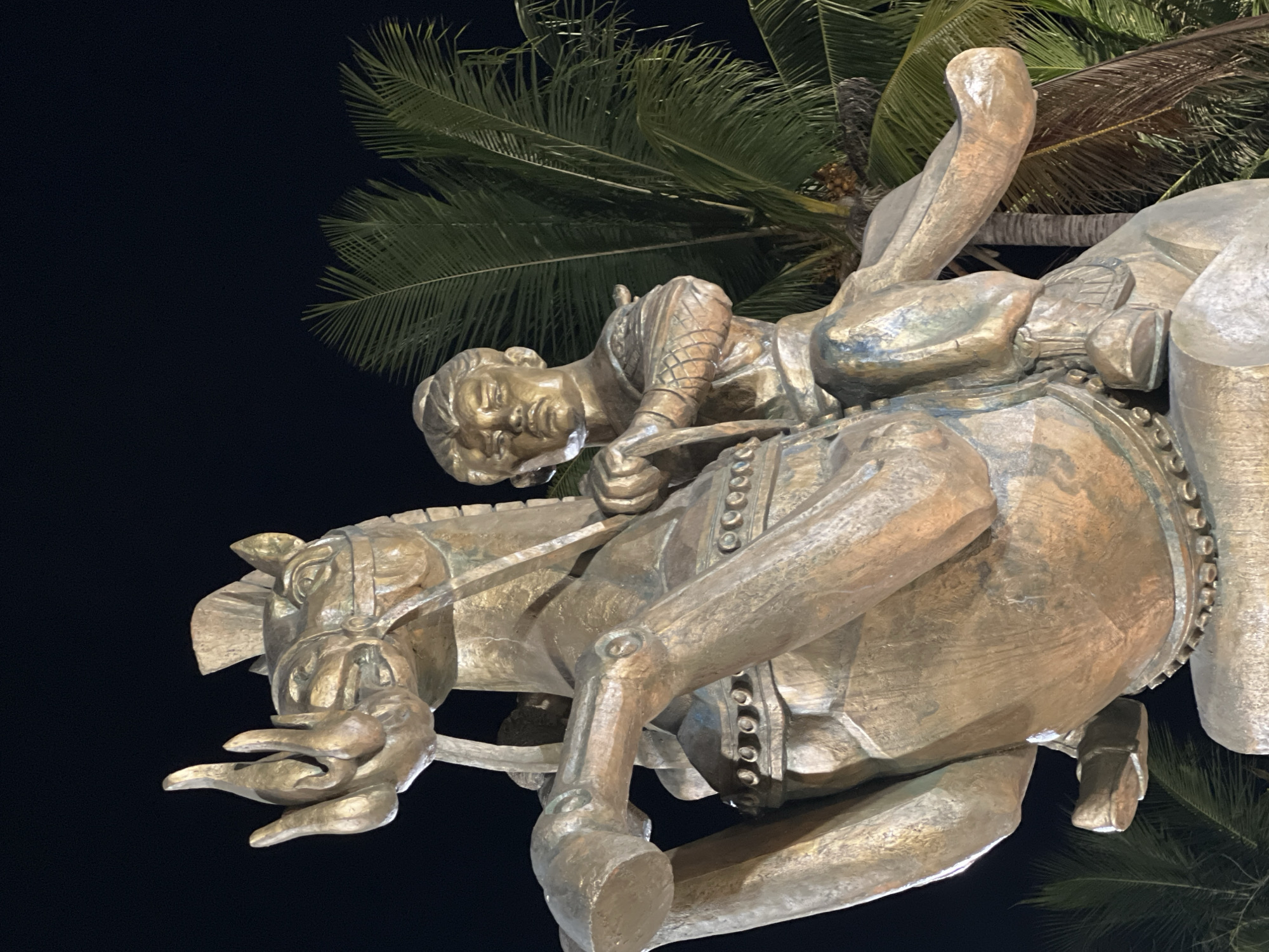 A bronze equestrian statue of a man is depicted against a dark night sky, with palm fronds visible in the background. The man, depicted with a stern expression and a warrior's attire, sits atop a horse that is rearing on its hind legs. The horse's mane appears stylized, with flames or similar elements emanating from its head. The statue is richly detailed, with intricate patterns on the man's armor and the horse's saddle. The sculpture is located in Nha Trang, Vietnam, specifically at the entrance to Vinpearl Land.