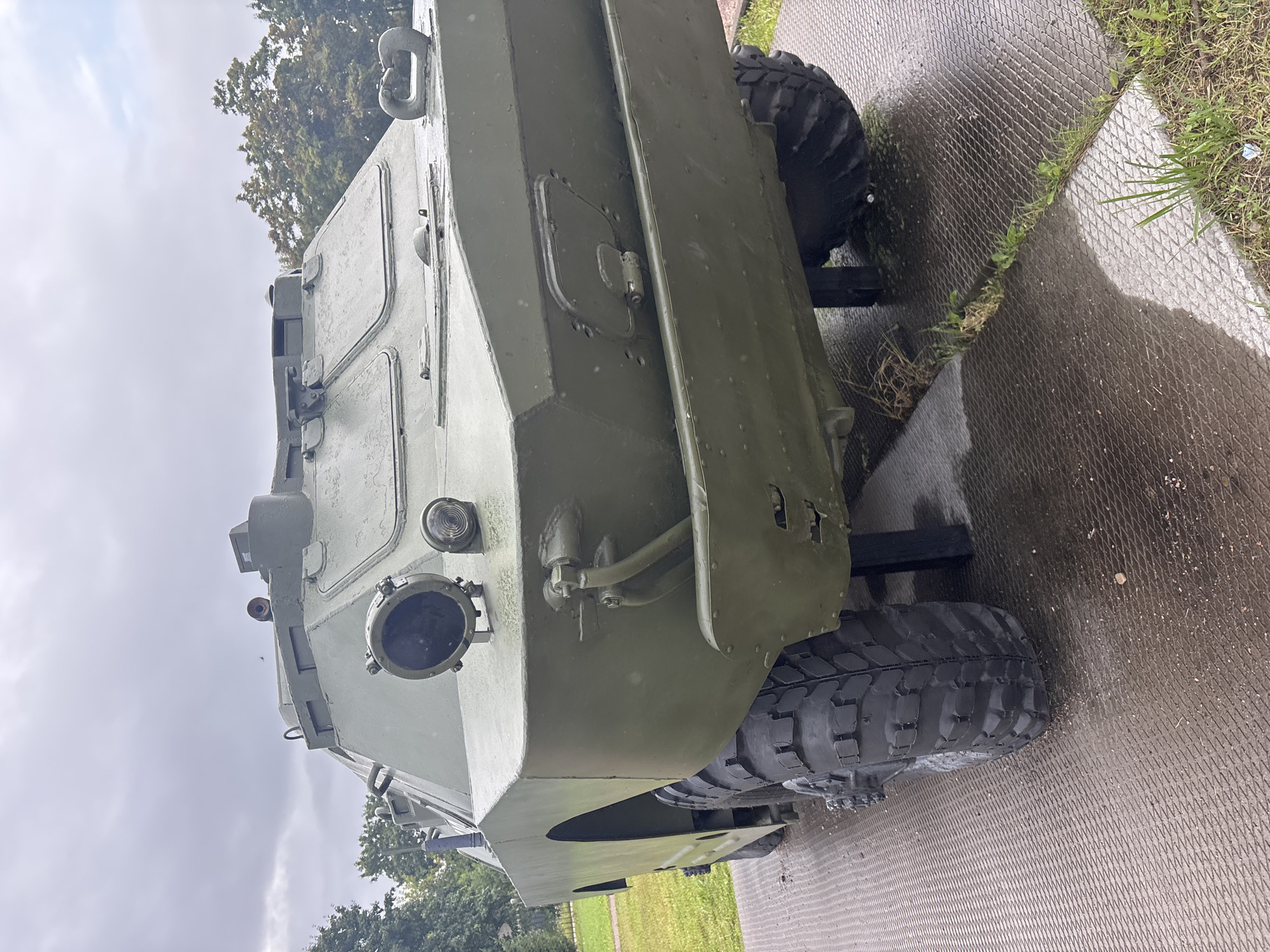 An olive-drab, wheeled armored vehicle is displayed outdoors on a wet, patterned metal platform. The vehicle is viewed from a low angle, from its front left side. It has large, rugged tires, a sloped front hull, and visible headlights and what appears to be a periscope or camera on the roof. The vehicle is green and shows signs of weathering. The ground is wet and reflects light. Beyond the vehicle, there is a grassy area and trees under a cloudy sky. The context of the location indicates this vehicle is a display at a museum or memorial in the city of Luga, Russia. There are no people visible in the image.