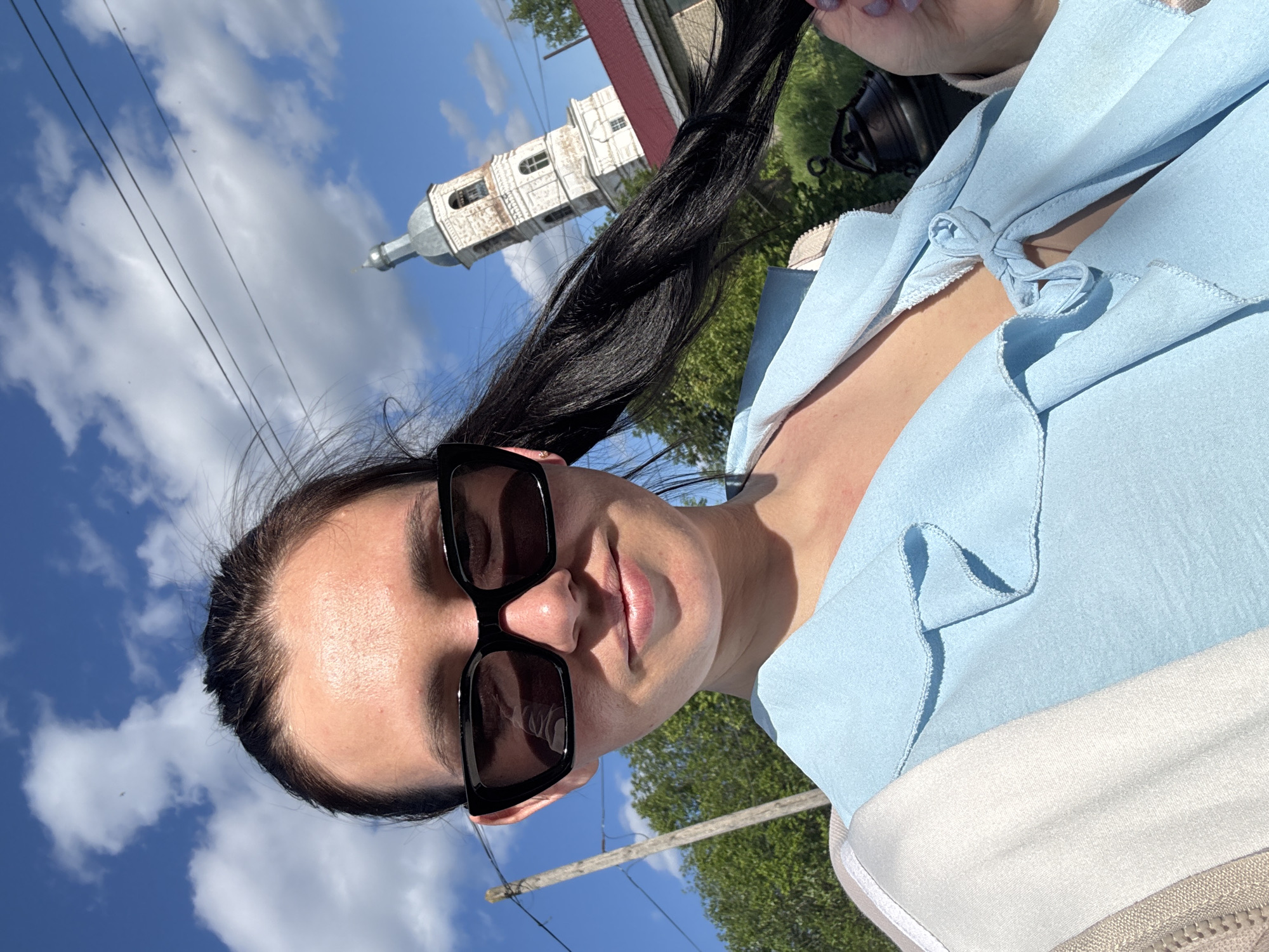An outdoor selfie captures a woman from the chest up under a bright blue sky with scattered white clouds, suggesting a clear and sunny day. The woman wears dark, rectangular sunglasses that obscure her eyes, and her lips show a slight, neutral smile. Her dark hair is pulled back. She is dressed in a light blue top featuring ruffles and a decorative knot tied at the neckline, possibly over a lighter, white or beige garment.
In the background, to the right of the woman, an old, light-colored architectural building, resembling a church or bell tower with distinct windows and a dome-like roof, stands against the sky. Green trees are visible in the mid-ground. Thin power lines stretch across the upper left portion of the sky, and a wooden utility pole is present in the lower background. The scene suggests a rural or small-town setting, fitting the provided context of Луптюг, Russia. No visible text is present in the image.