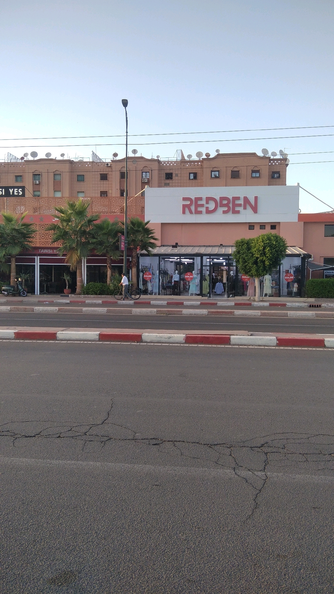 The image depicts a street scene in Marrakesh, Morocco, featuring a commercial building with a prominent red "REDBEN" sign. The building appears to house clothing stores, with mannequins displaying garments visible through glass storefronts. Signs reading "SOLDE" (sale) are also visible in the shop windows. To the left, another store is labeled "TAWSI YES." A person is seen riding a bicycle on the sidewalk in front of the shops, and a scooter is parked nearby. The street in the foreground shows signs of wear, with visible cracks in the asphalt. Palm trees are planted along the sidewalk, and satellite dishes are mounted on the rooftop of the adjacent building. The sky is clear and blue, suggesting it is daytime.
