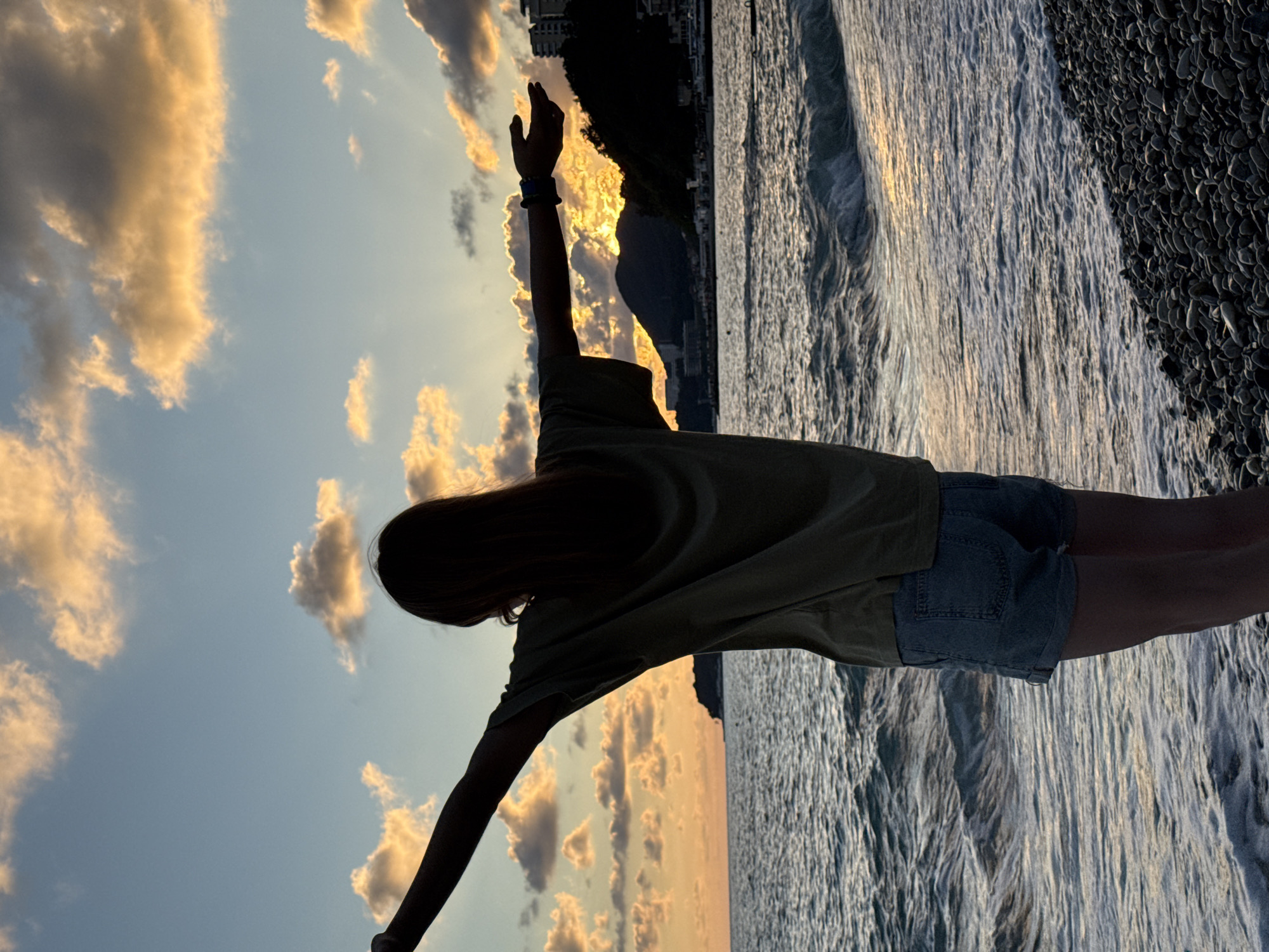 A person stands with their arms outstretched facing a sunset over the sea on a pebbled beach in Nebug, Russia. The sky is filled with clouds illuminated by the setting sun, casting a warm glow. The sea has moderate waves breaking on the shore. In the background, there is a dark silhouette of a hillside with buildings. The person is silhouetted, wearing a t-shirt and denim shorts.