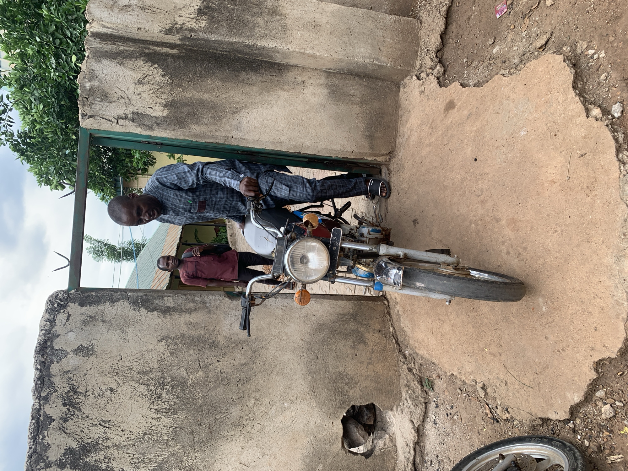 The scene is a concrete entranceway in Akwanga, Nigeria, possibly leading to a residence or building. An adult male wearing a gray plaid suit and black sandals is seated on a vintage motorcycle, which is positioned inside the entranceway. Another adult male is standing in the background, wearing a maroon shirt and carrying a dark bag over his shoulder. The immediate surrounding environment consists of aged concrete structures, a green metal gate, and exposed earth. The overall condition of the area suggests a state of disrepair. A spare tire is on the ground near the edge of the picture.