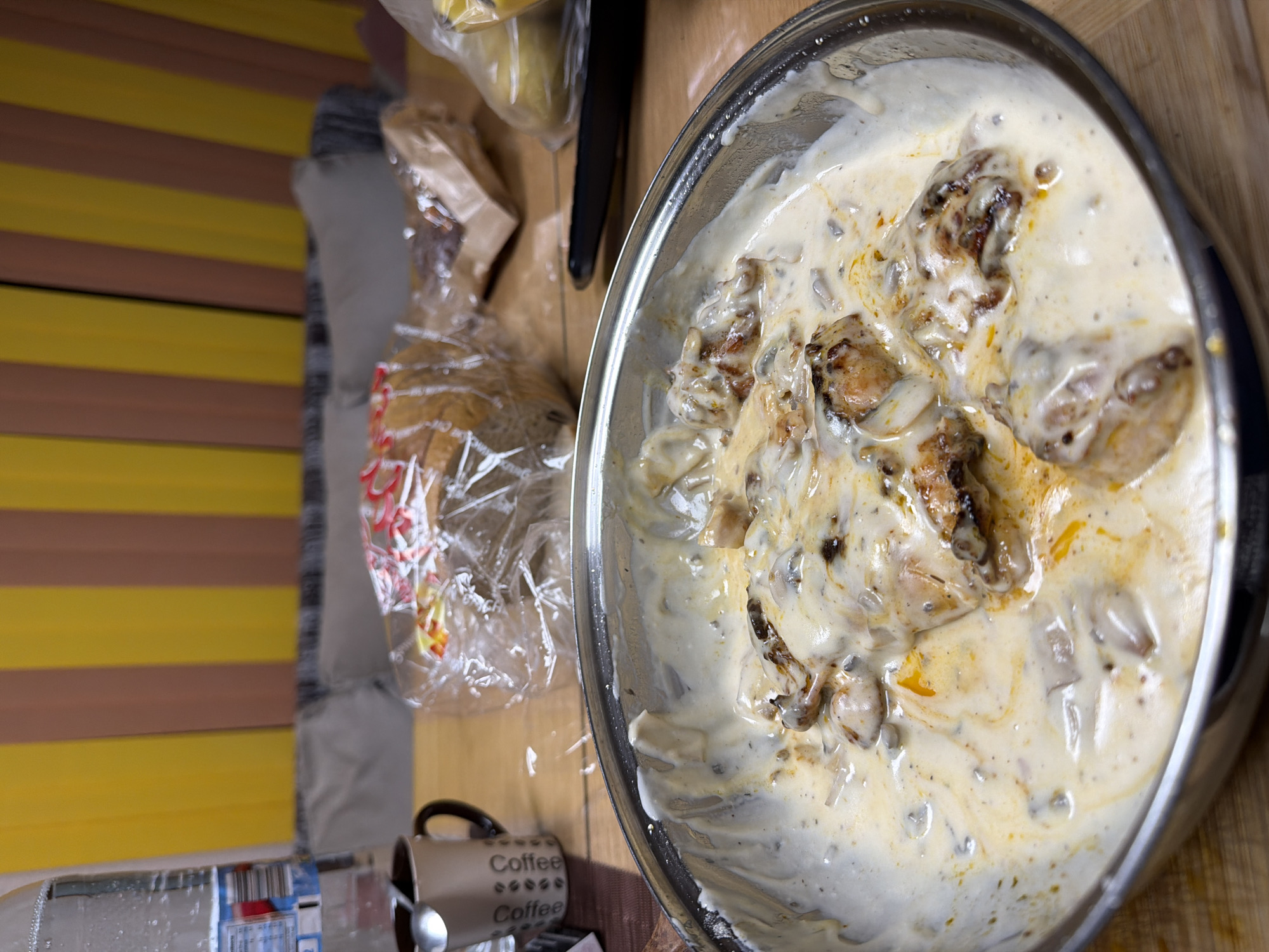 A close-up, high-angle shot displays a metal mixing bowl filled with a creamy white sauce interspersed with pieces of what appear to be chicken or another type of meat. The sauce has a speckled appearance, possibly due to the inclusion of herbs or spices, and some areas are tinged with a yellowish or orange hue, potentially from oil or fat.
To the left of the bowl, a plastic-wrapped loaf of bread sits on a wooden surface. Behind the bread, a brown paper bag and some items wrapped in clear plastic are also visible. In the lower left corner of the frame, there is a partially visible water bottle and a mug labeled "Coffee" multiple times. A spoon rests next to the mug.
The backdrop includes a striped pattern of yellow and brown, potentially indicating a curtain or wall covering.
The location and city of Panagyurishte, Bulgaria is not determinable from the objects in the image.