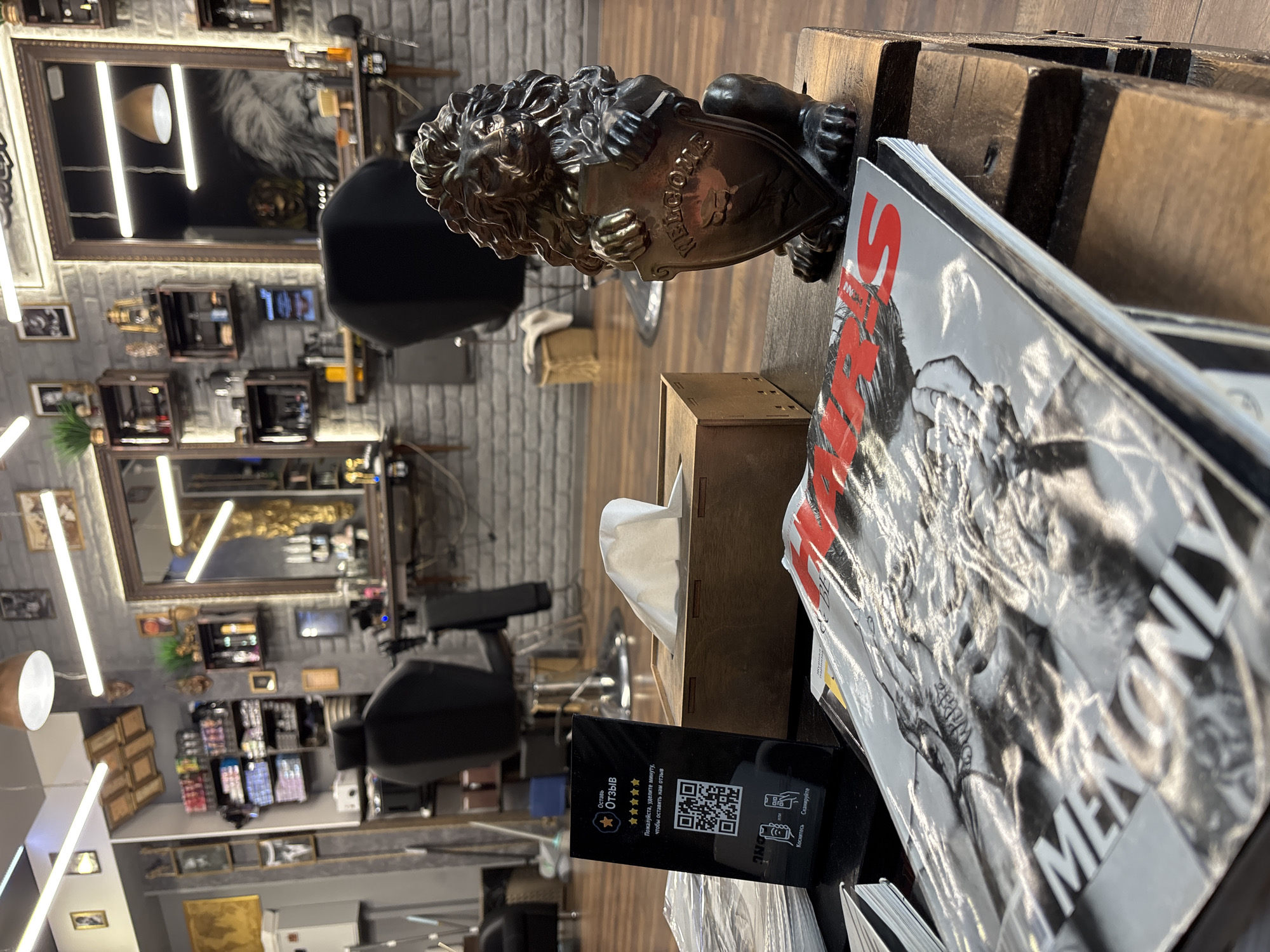 The image depicts the interior of what appears to be a barbershop, likely in Minsk, Belarus, based on Cyrillic text visible. A lion statuette and a stack of "MEN ONLY" magazines sit on a wooden stand with a tissue box and a black display sign with a QR code and text in Cyrillic, encouraging customers to leave a review. In the background, barber chairs, mirrors, product displays, and framed pictures are visible, indicative of a barbershop or salon environment.