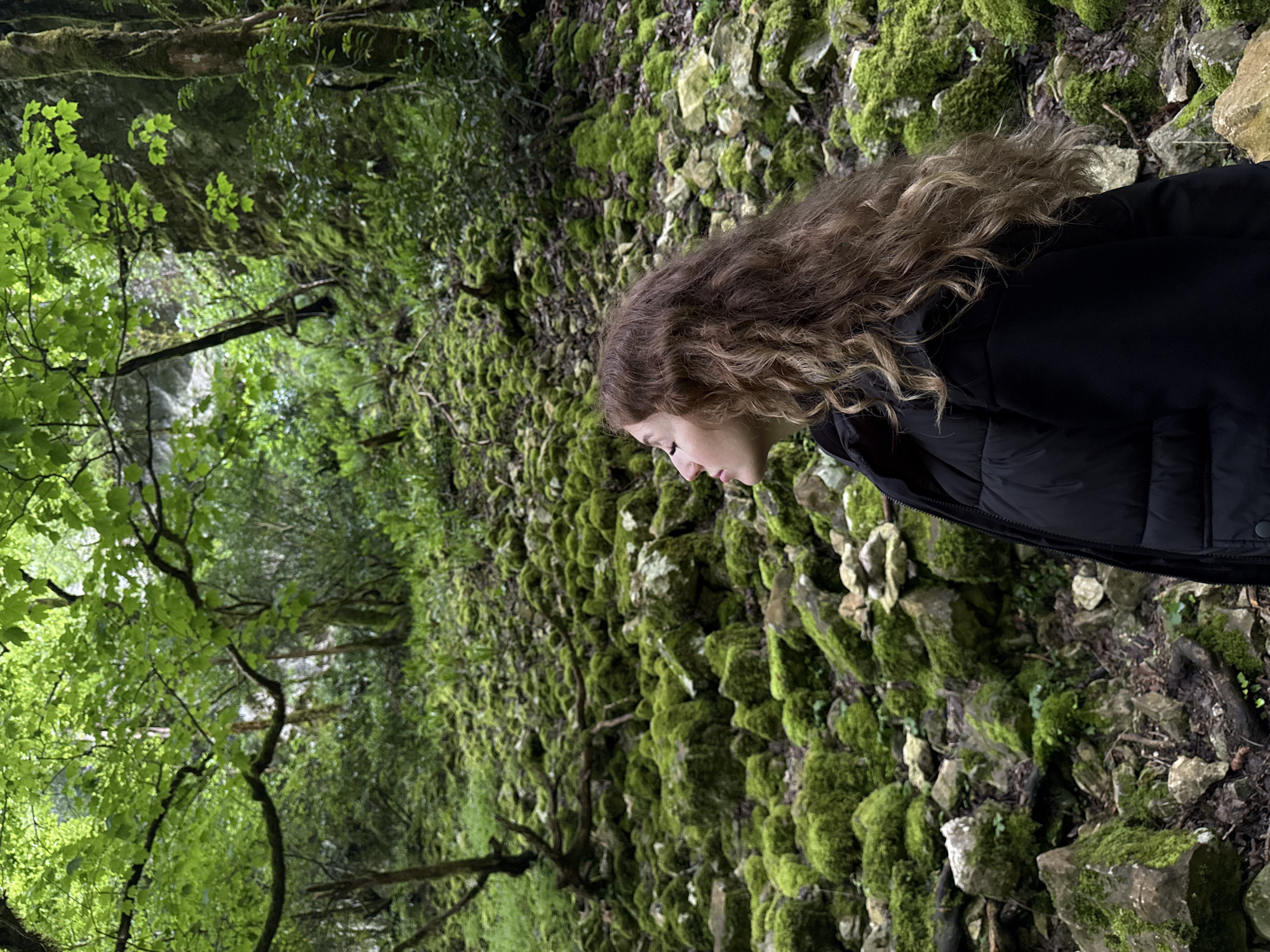 A person with wavy brown hair is standing in a forest in Tsabal, Abkhazia. The person is wearing a black jacket. The forest is characterized by moss-covered rocks and lush green foliage. The camera angle is rotated counter-clockwise, so the scene appears tilted.