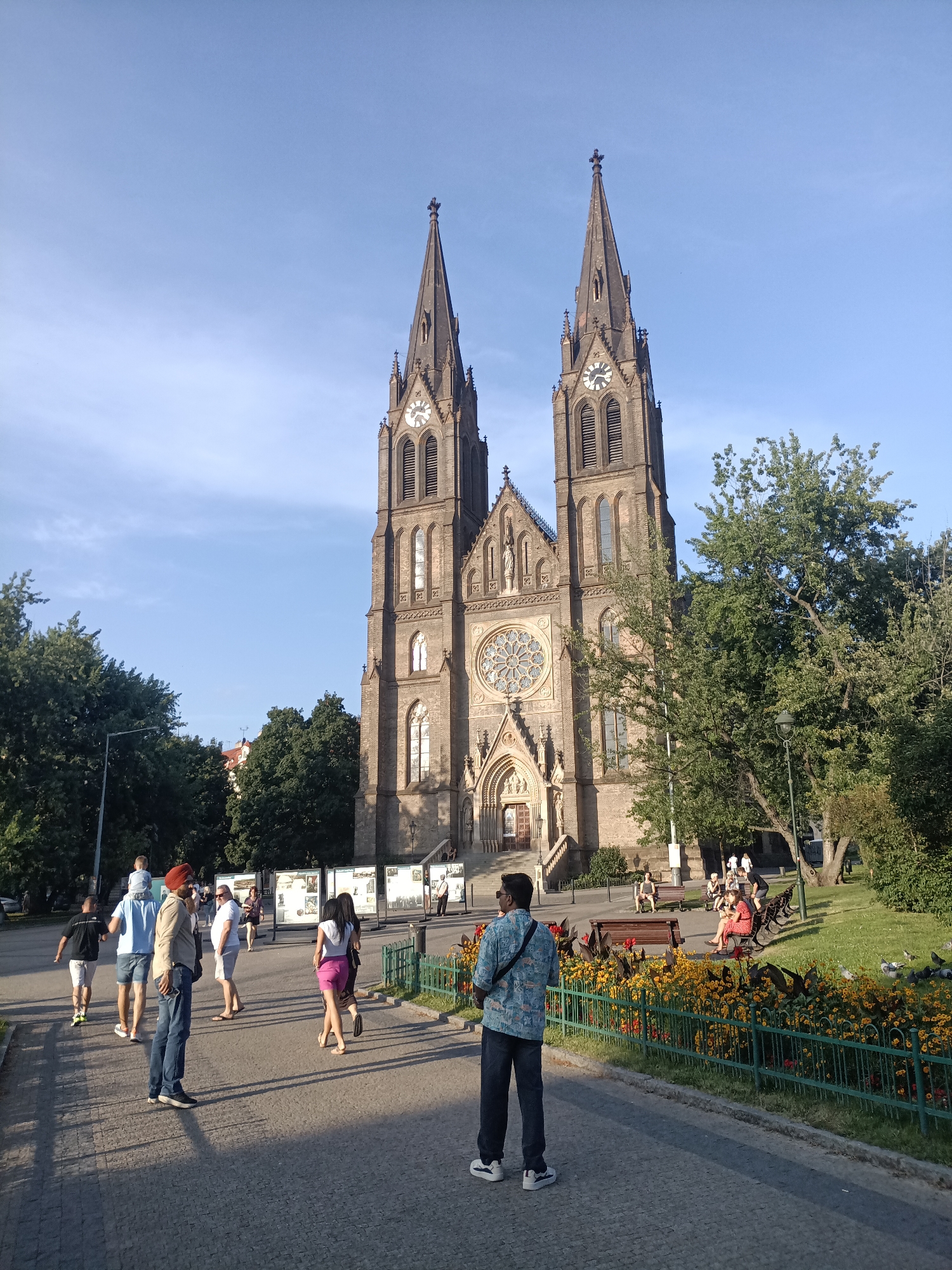 This image depicts the exterior of the Basilica of St. Peter and St. Paul in Vyšehrad, Prague, Czechia. The grand Gothic Revival basilica features two prominent spires, a large rose window above the entrance, and intricate architectural details.
In the foreground, several people are visible on a paved walkway. A man wearing a blue shirt and jeans stands facing away from the camera, looking towards the basilica. To his left, a man in a light blue shirt and denim shorts walks with a child on his shoulders, accompanied by another man in a black shirt. Further to the left, a man wearing a turban and a light brown shirt is walking with his back to the camera.
To the right of the main walkway, a manicured green lawn is dotted with benches occupied by several individuals. A vibrant flowerbed with yellow and red flowers borders the lawn, separated by a green metal fence. Trees provide shade in the background, and a lamp post is visible on the left side of the image. The sky is clear and blue, indicating a sunny day. Several display boards with images are set up on the left side of the walkway, suggesting an exhibition or informational display.