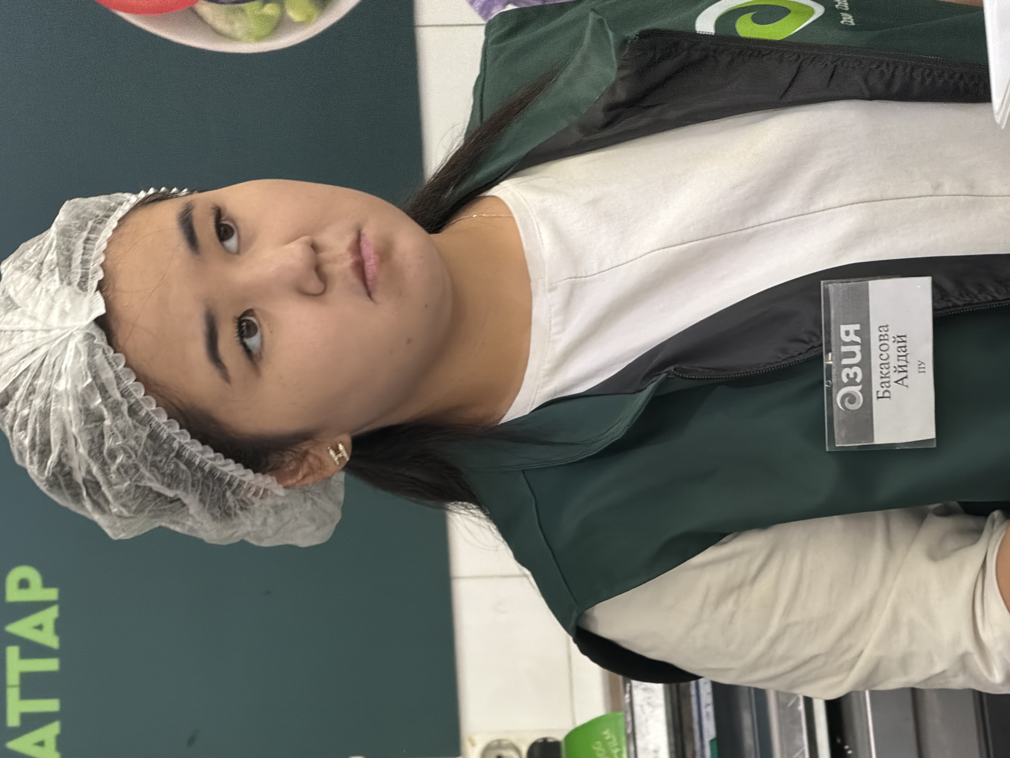 A young woman with dark hair wears a hairnet, a white t-shirt, and a dark green vest. She has a name tag on her vest that reads "АЗИЯ" and "Бакасова АйДай" and "ЛУ". She is standing in what appears to be a market or store, with a dark green wall behind her. The woman is looking to her right.