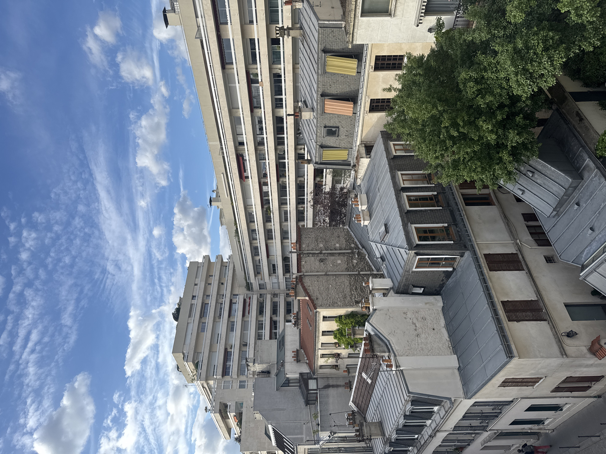 An angled, high-level outdoor shot captures a slice of urban life in Paris, France. The scene is dominated by buildings of varying heights and styles. A tall, modern apartment building with balconies and linear facade is on the left. To the right, a cluster of older, traditional Parisian buildings with zinc roofs, dormer windows, and shutters. A tree adds a touch of greenery. The sky is a bright blue with streaks of clouds. In the bottom right corner, a glimpse of people walking along a street.