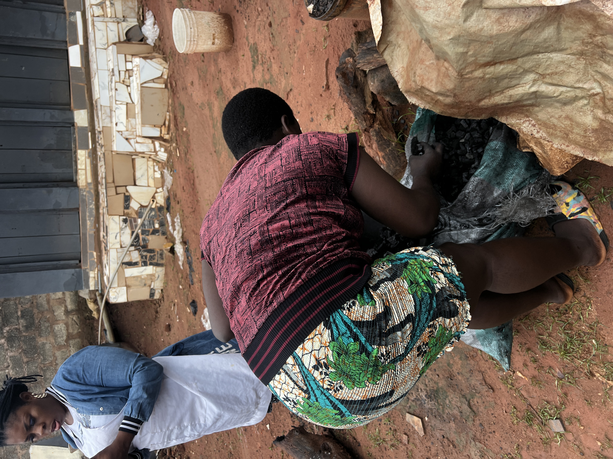 In Nkpor, Nigeria, a woman is shown bending down and reaching into a large, open bag. She is wearing a patterned skirt with green, blue, and white colors, and a maroon top with a black and red striped band around the waist. Her hair is styled in braids. Beside her, another woman in a white apron and blue denim jacket looks down at a phone in her hand. The ground is covered in red dirt and scattered with small green plants. In the background, a building with a tiled facade and a dark metal door is visible, along with a white bucket. The scene appears to be an outdoor, possibly domestic or marketplace setting.