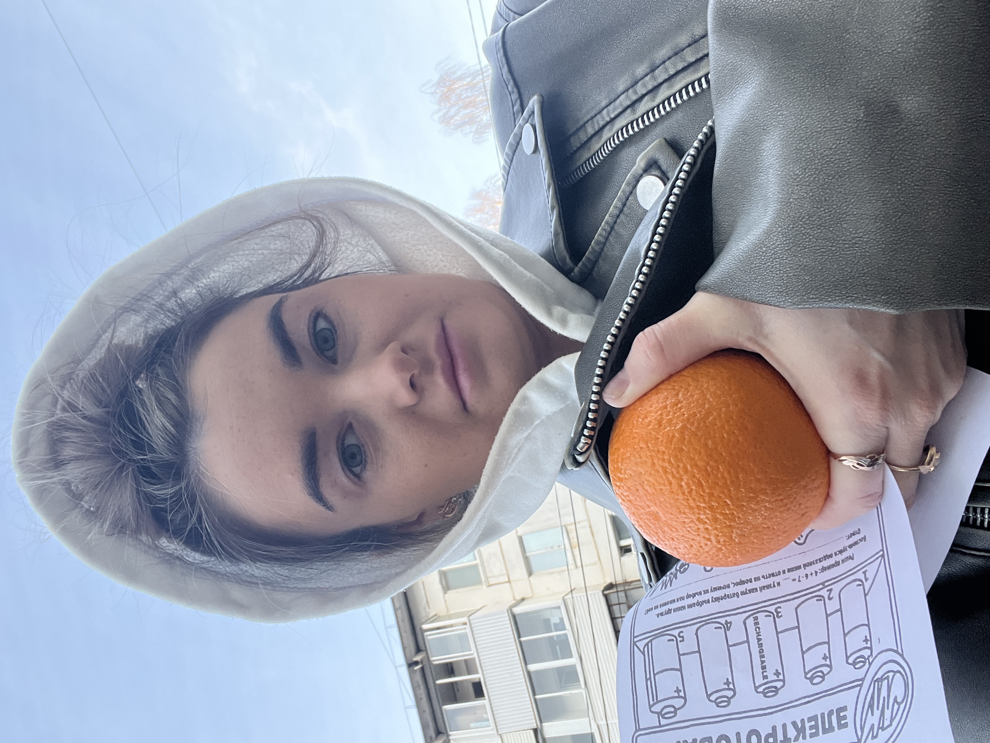 A woman wearing a white hooded sweatshirt and a gray leather jacket is holding an orange and a piece of paper with a diagram of batteries. The paper has text in Russian: "ЭЛЕКТРОТовм" and "Реши пример: 4+6-7=". In the background is a multi-story building with windows. The image was taken from a low angle, looking up at the woman and the building. The location is the city of Chaikovsky, Russia.