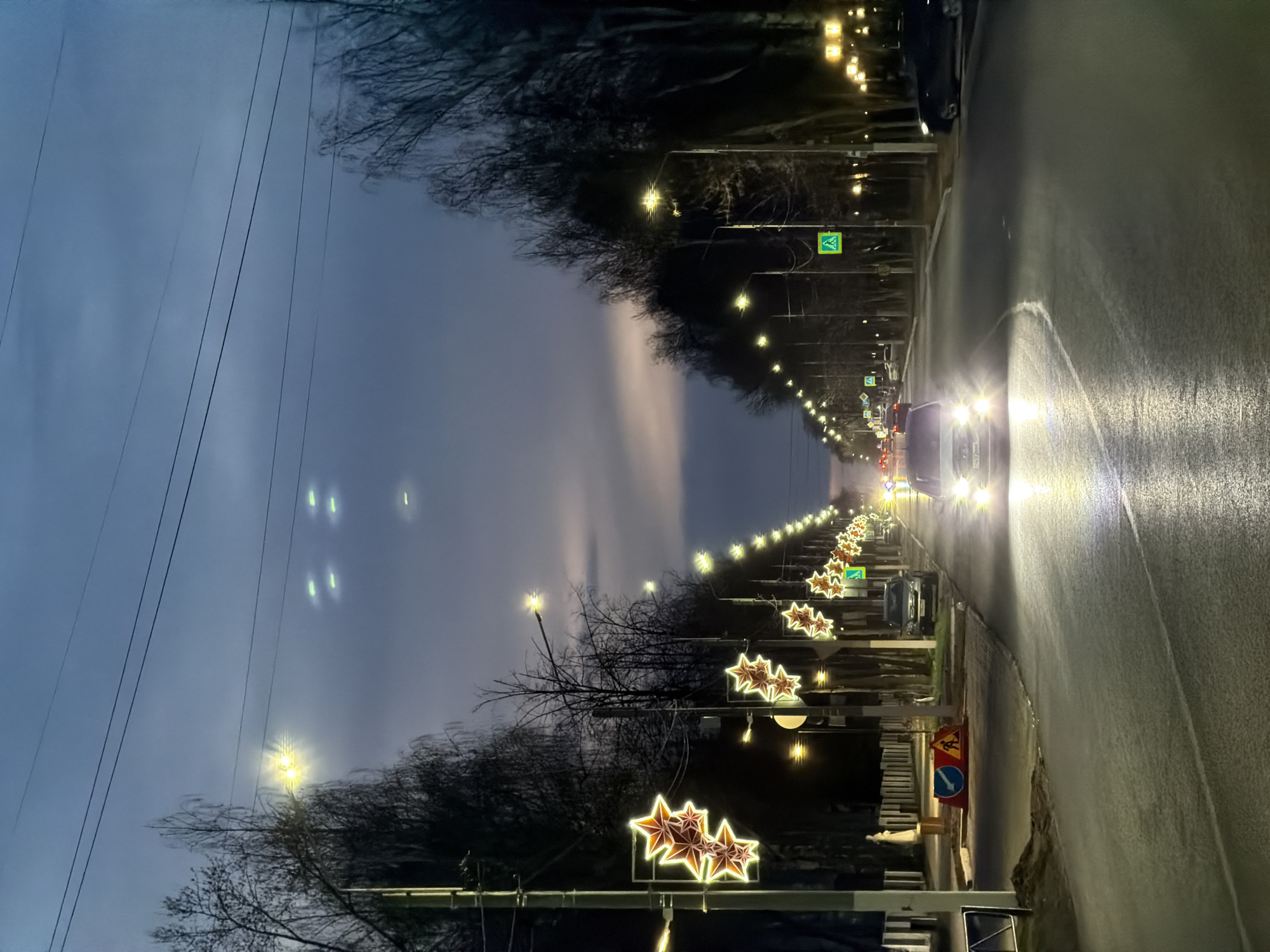 This image captures a long, straight road in Chaikovsky, Russia, during what appears to be nighttime or late dusk. The road, likely asphalt and appearing wet from rain or melted snow, recedes into the distance, lined on both sides by trees with bare branches, suggesting a colder season like late autumn or winter.
Numerous streetlights illuminate the scene, casting a warm glow along the thoroughfare. On the left side of the road, decorative star-shaped lights, possibly part of holiday or festive decorations, are visible on poles. Several vehicles are present; one car in the foreground on the right has bright headlights shining towards the viewer, and other vehicle lights are visible further down the road. Utility wires span across the cloudy, dark blue-grey sky overhead.
Notable details include:
- **Road signs:** Green square pedestrian crossing signs with white downward arrows are visible on both sides of the road. On the right side in the foreground, there's a yellow triangular warning sign depicting figures, likely indicating roadworks, and a blue circular sign with a white arrow pointing right, indicating a mandatory turn direction.
- **Lighting:** The streetlights, car headlights, and decorative stars provide significant illumination, creating reflections on the wet road surface.
The scene depicts regular urban or suburban street activity during an evening, possibly around a festive period given the star decorations. No people are clearly visible, and no specific event beyond typical traffic flow is apparent. No legible text is visible on signs or vehicles.