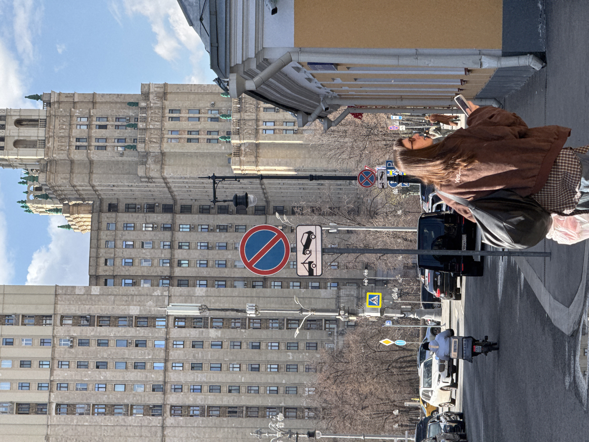 The scene is set in Moscow, Russia, and features a street with a prominent Soviet-era skyscraper in the background. A woman is looking at her mobile phone and holding it with her right hand. She is wearing a brown jacket, a plaid skirt, and a black shoulder bag. Several road signs are visible, including "No Parking" and "Pedestrian Crossing." Various cars are parked along the street, and a motorcyclist is driving down the road. The weather appears to be sunny. The architecture of the buildings indicates a post-war Soviet style.
