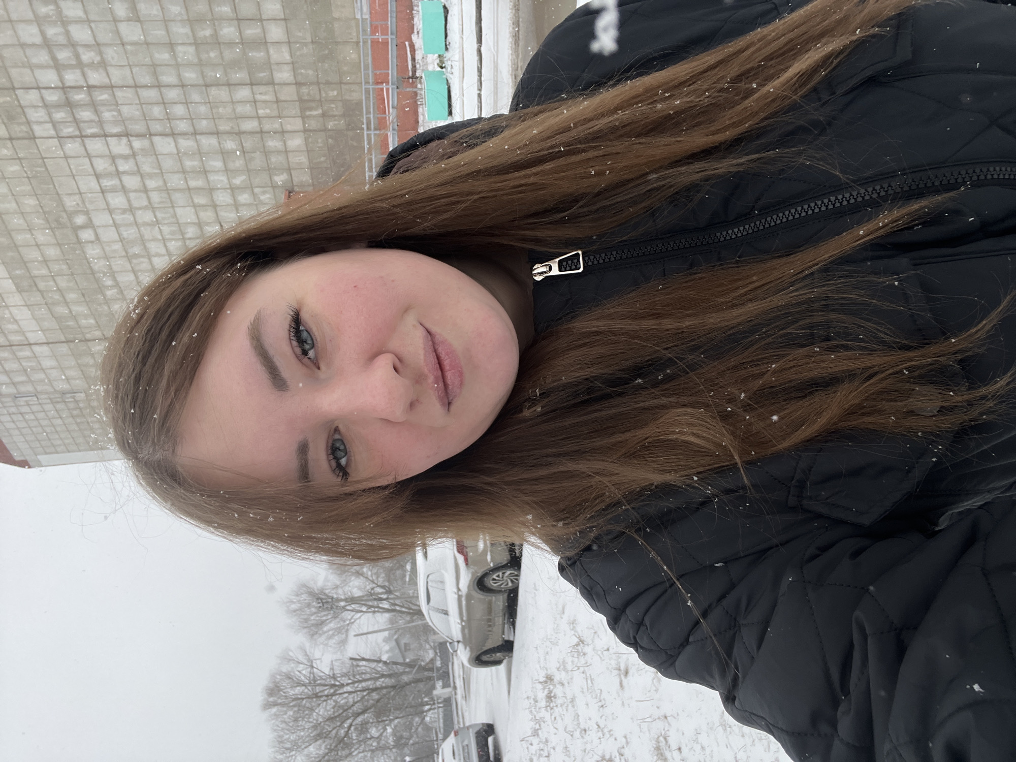A woman with long brown hair and blue eyes is wearing a black puffer jacket outdoors in snowy conditions. Snowflakes are visible on her hair and clothing. In the background, there is a snow-covered parking lot with cars and bare trees. The building in the background appears to be a multi-story structure with light-colored brick or tile. Given the prompt, it's inferred the location is Chaikovsky, Russia.