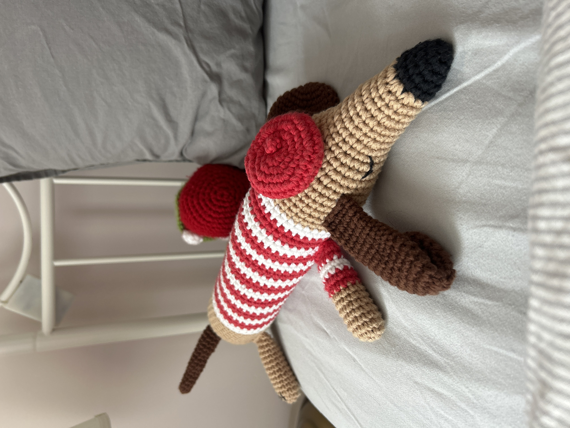 A crocheted toy dachshund wearing a red and white striped sweater and a red beret is lying on a gray bedsheet. The dog has a tan body, brown ears and paws, and a black nose. A red and green crocheted object is near its rear. A white metal bed frame is visible. The location is unknown and cannot be determined from the image.
