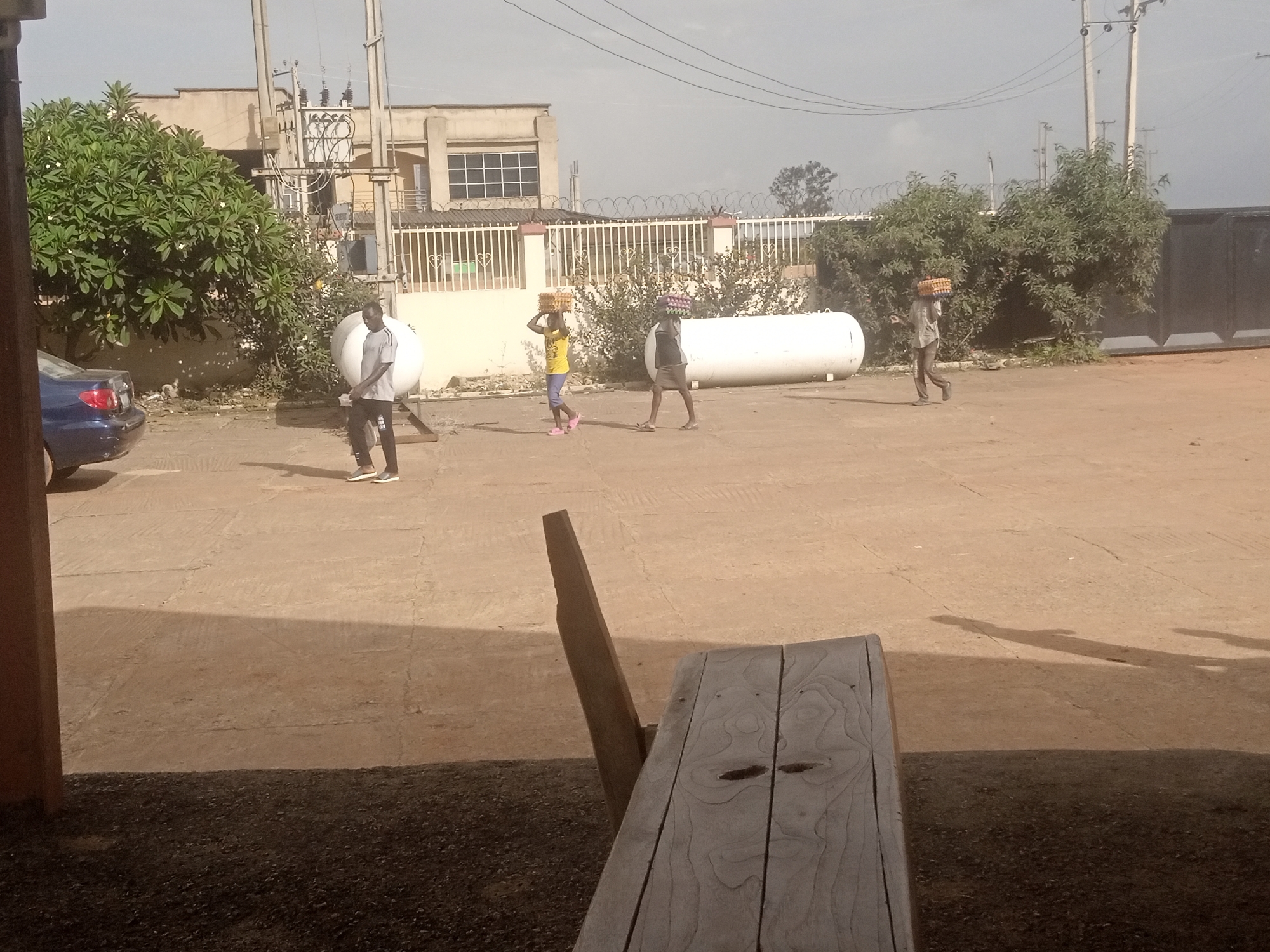 The image depicts an outdoor scene with several people carrying loads on their heads. In the foreground, a wooden picnic table and bench are partially visible, casting a shadow on the ground. The background shows a building with a white fence, a large white cylindrical object that appears to be a tank, and some trees. Overhead, power lines and utility poles are present.
Four individuals are visible walking across a paved area. One man, wearing a grey t-shirt and dark pants, is walking away from the camera towards the left. Further to the right, a woman in a yellow shirt and blue pants is carrying a yellow basket on her head, with a person in a dark shirt and skirt also carrying a similar basket on their head. To the far right, another person in a dark shirt and pants is walking away from the camera, also carrying a basket. The loads appear to be stacks of goods, possibly food items.
The scene appears to be set during the daytime, with the sun casting distinct shadows, suggesting it's likely morning or late afternoon. The weather seems fair, with a hazy sky. The presence of the building, tank, and utility poles suggests an industrial or commercial area, possibly a market or distribution center. There is no visible text in the image. The overall mood is one of labor and movement.
