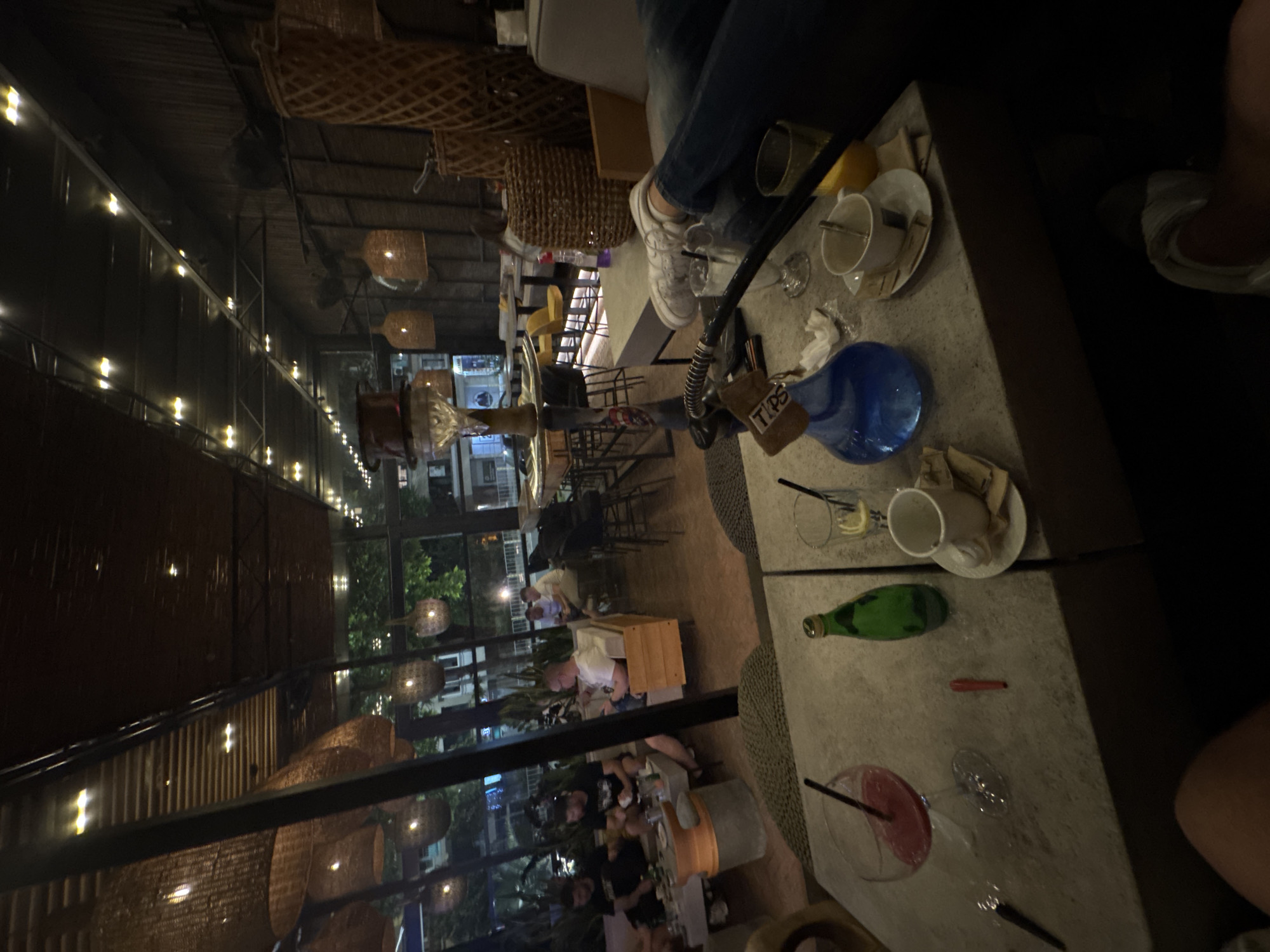 The image shows the interior of a cafe or lounge in Burgas, Bulgaria, likely at night. Visible through the large windows are urban street scenes with trees and buildings, illuminated by streetlights. Inside, the ceiling is decorated with woven basket-like light fixtures and fairy lights. Several tables and chairs are set up, and patrons are seated at some tables.
A hookah sits on the table, along with several glasses, cups, plates, and a bottle. People's legs and feet can be seen. The scene suggests a relaxed social gathering.