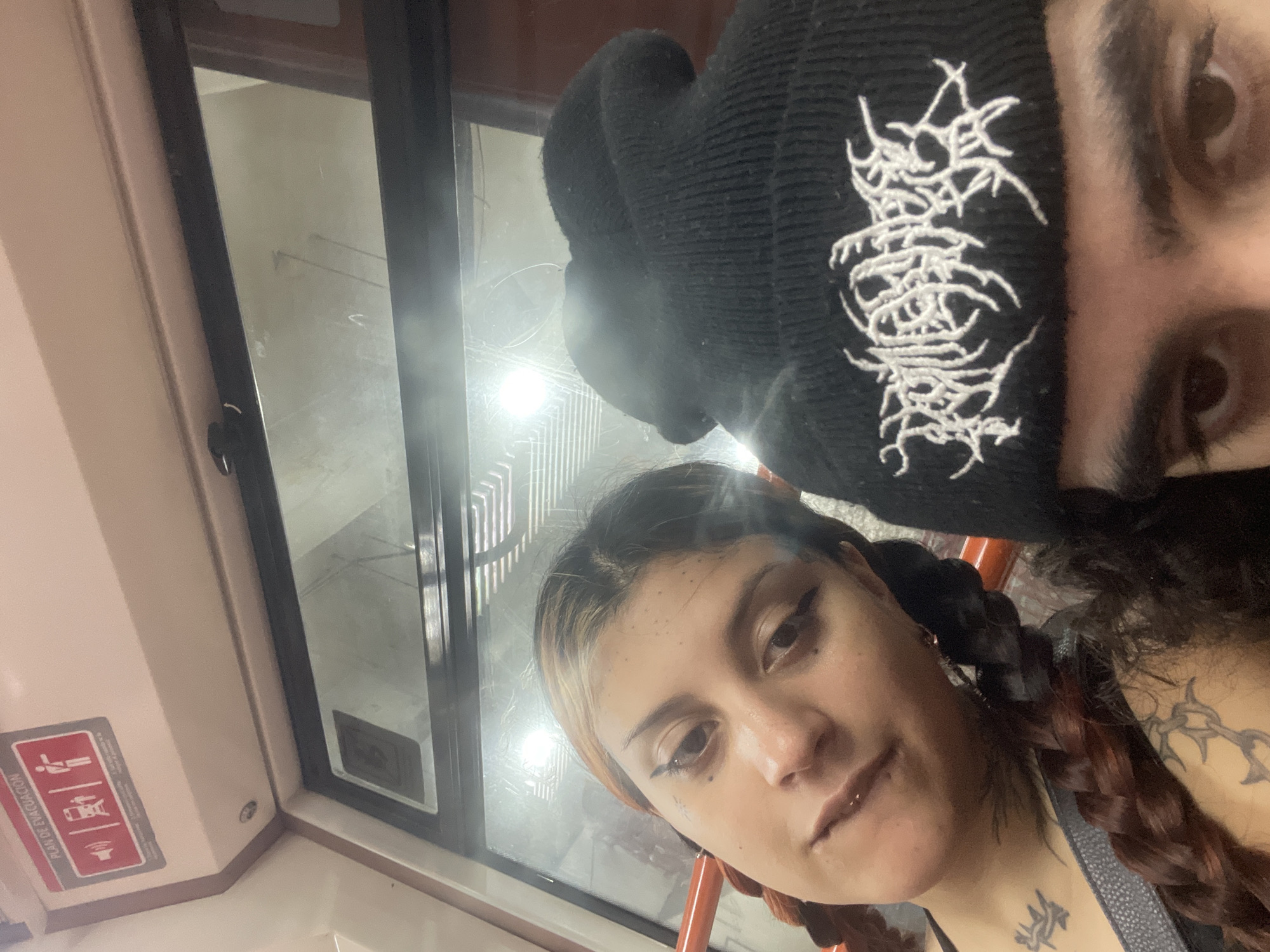 Two individuals are depicted in the frame. One individual, positioned above and slightly to the right, wears a black beanie with intricate white embroidery. The beanie obscures a portion of their face, revealing an eye and eyebrow. The second individual, located below and to the left, is a person with light brown hair with blonde streaks in braids. They are wearing eye makeup and a lip gloss. Both individuals appear to be within a public transportation vehicle, evident by the window and the evacuation plan signs. The location is suggested to be the city of Buenos Aires, Argentina, based on the signage and vehicle design.
